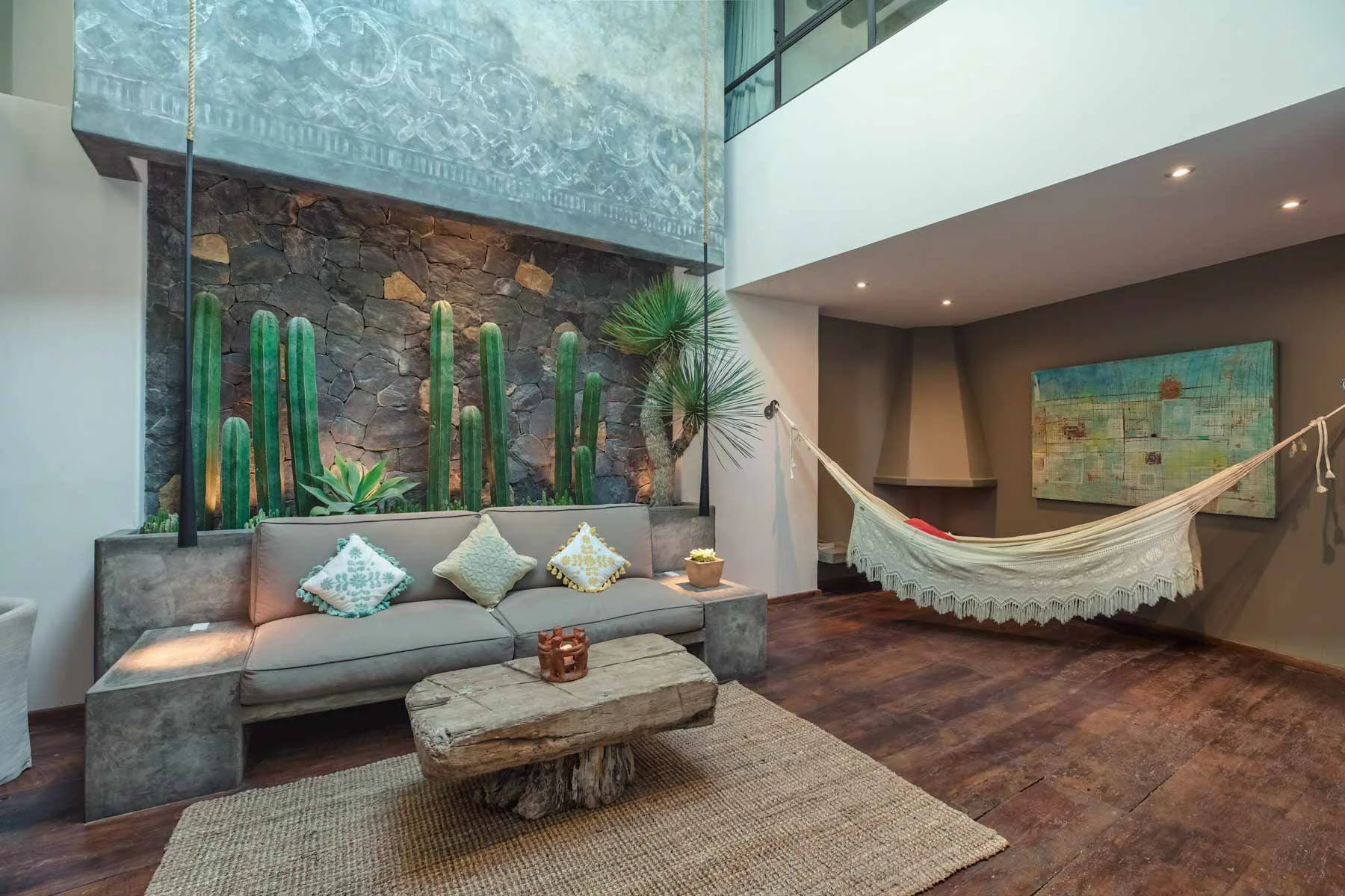 A modern living room with a concrete sofa decorated with patterned throw pillows, a rustic wooden coffee table, a beige rug, tall green cacti and plants against a stone wall, a light-colored hammock secured to the ceiling, and a colorful abstract pai