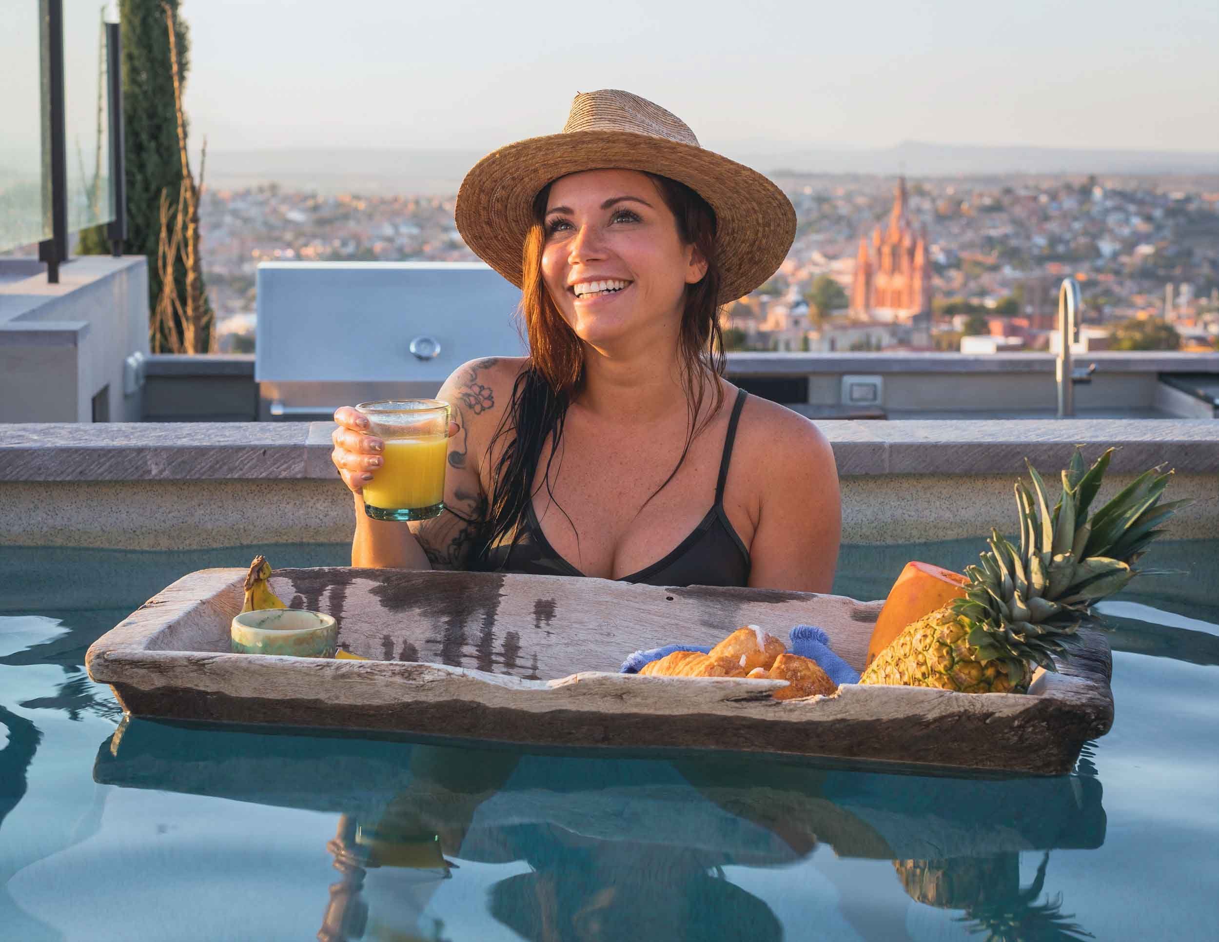 Woman smiling, wearing a straw hat and a black swimsuit, holding a glass of orange juice, sitting by a pool with a city skyline and church in the background, with fruits and a wooden tray on the pool edge.
