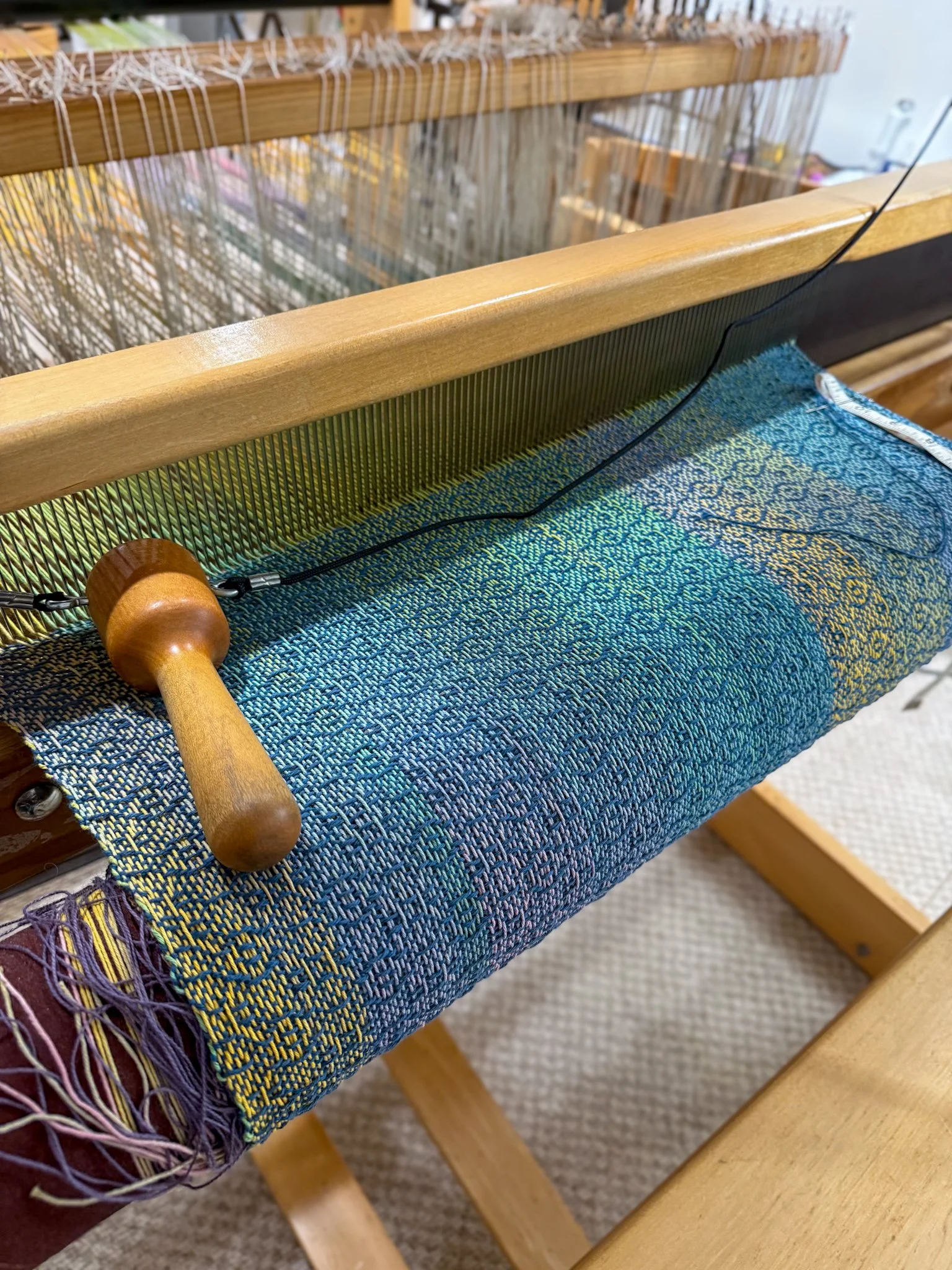 A close-up of a wooden loom with colorful woven fabric and a black cord. The loom's wood is smooth and light-colored, with a rounded handle visible at the bottom left corner. There are threads and weaving materials on the fabric, which features an in