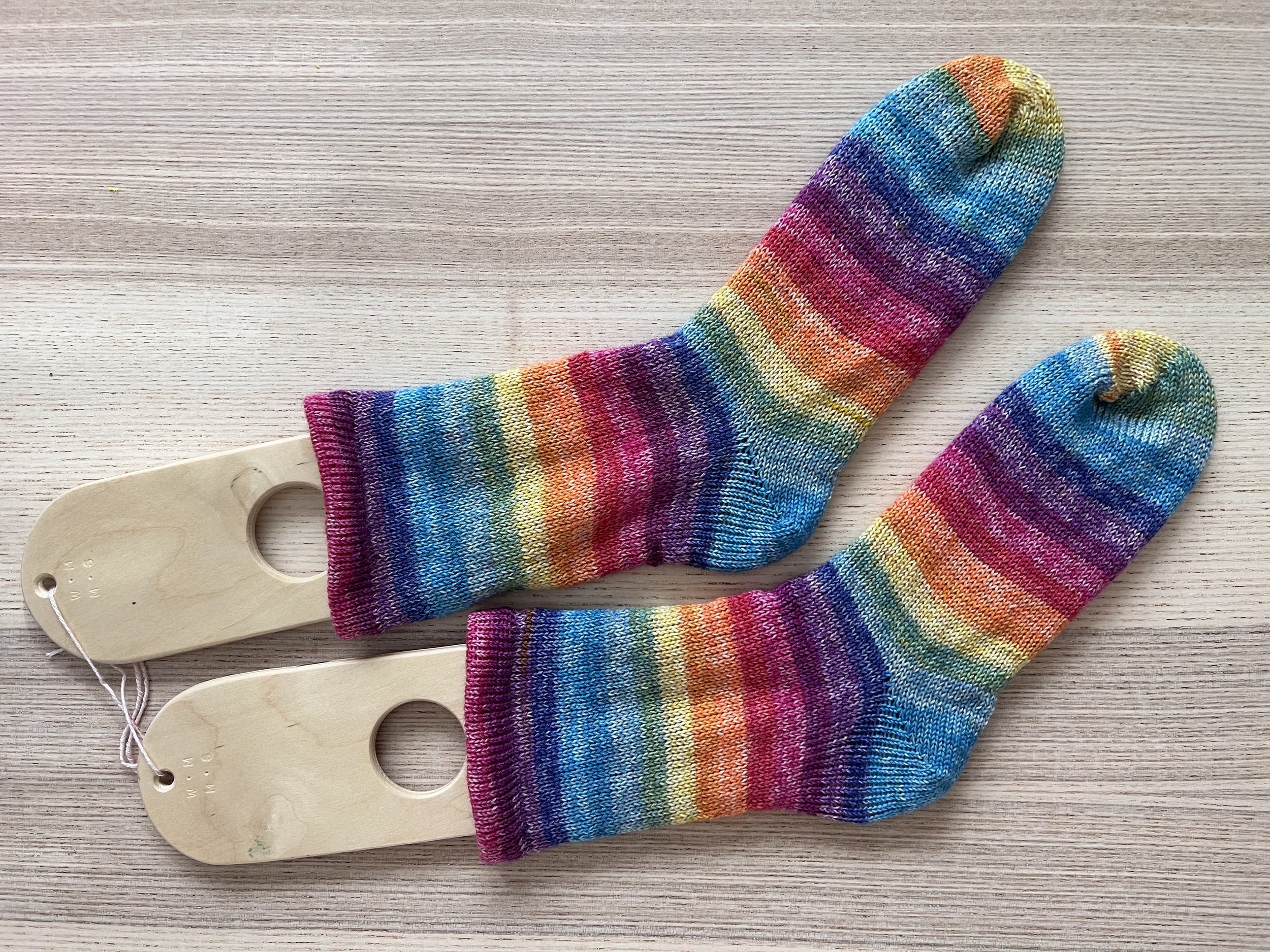 Pair of rainbow-colored striped socks with wooden sock blockers on a light wooden surface.