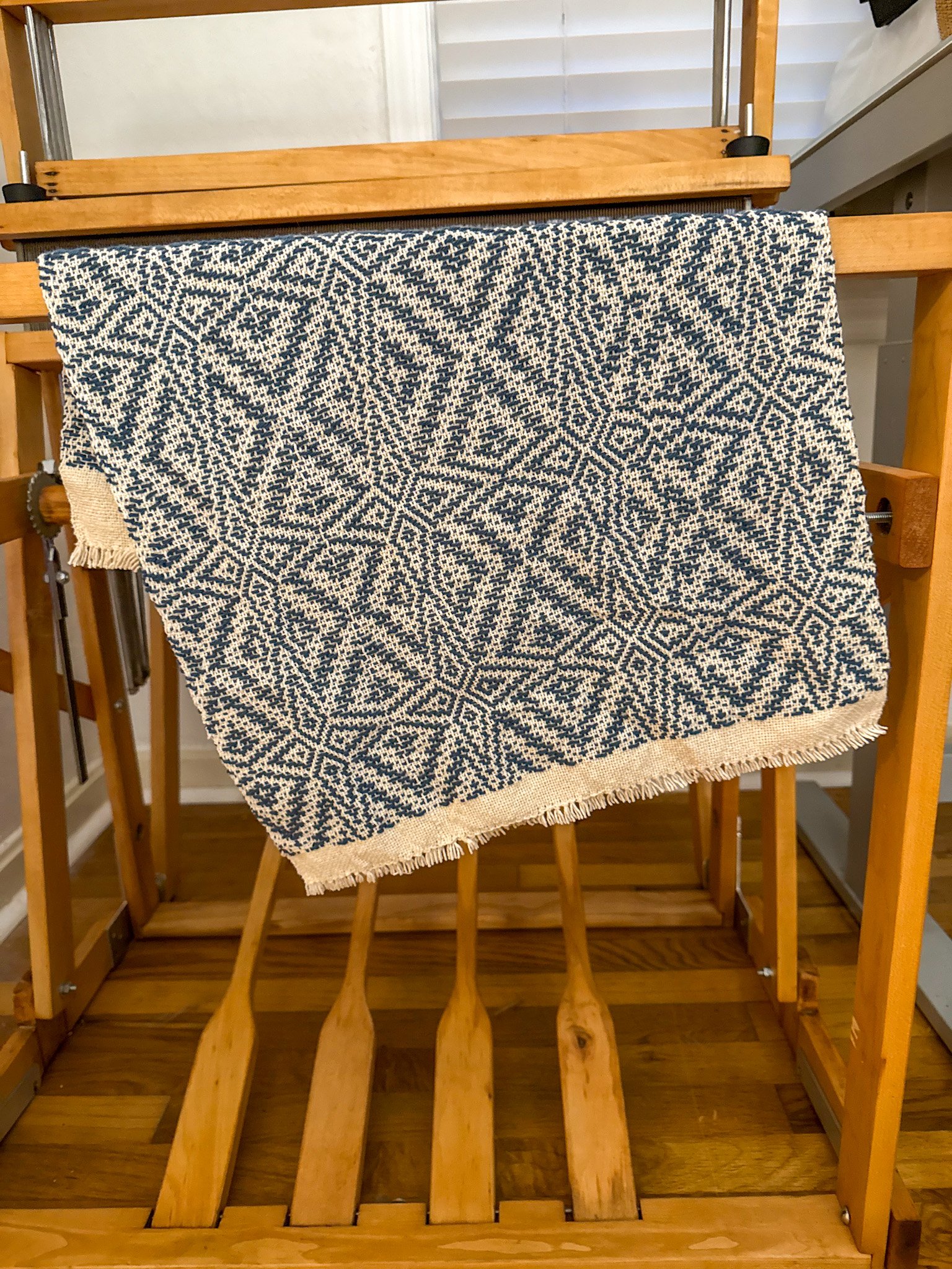 A loom with a hand woven tea towel draped over it, standing on a hardwood floor in front of a window with closed blinds.