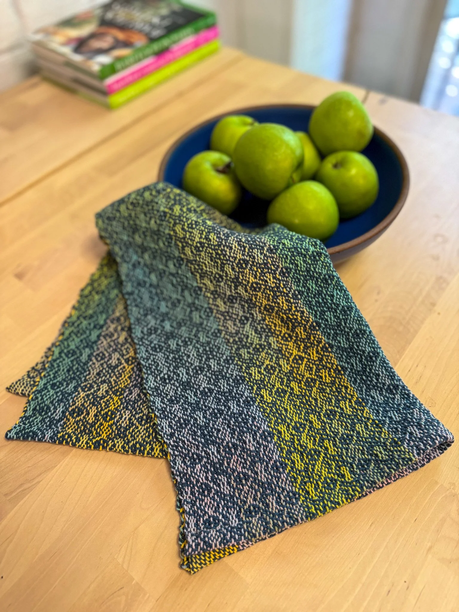 A flat-lay photograph of a multicolored woven tea towel with a yellow, black, and pink pattern, placed on a wooden surface. In the background, there is a blue bowl filled with green apples and a stack of magazines.