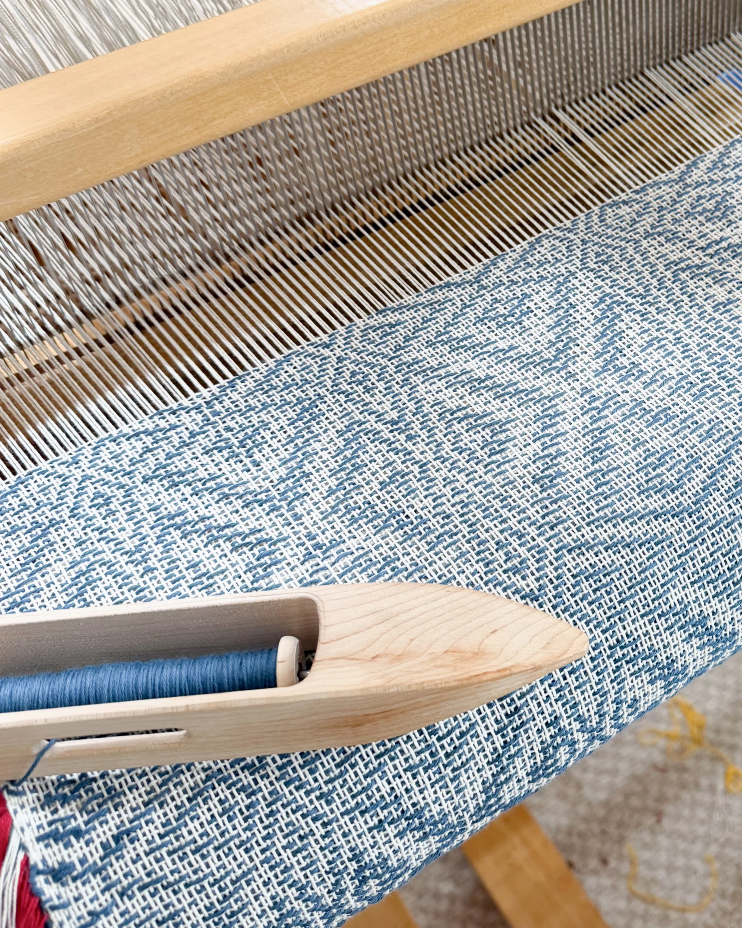 Close-up photo of a weaving loom with blue and white woven fabric, with a wooden shuttle placed on the fabric.