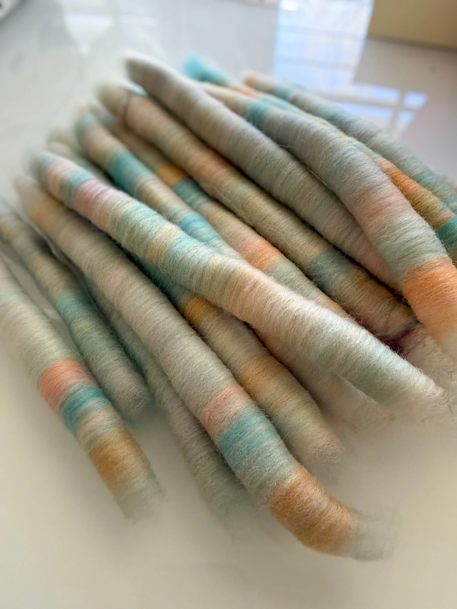Close-up of hand carded wool rolags with pastel colors, purple, blue, green, orange, and beige, on a white surface.
