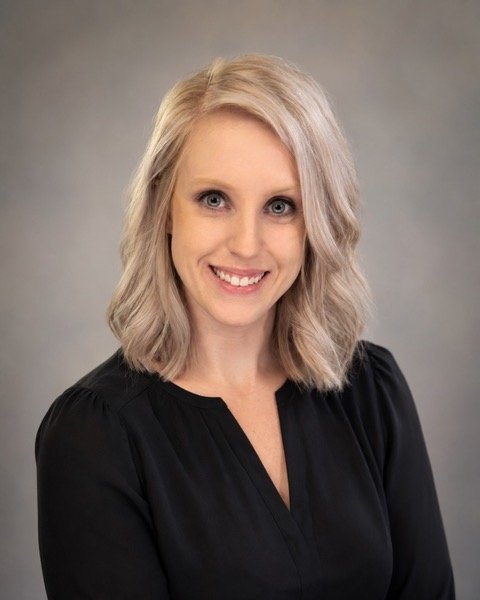 A woman with shoulder-length blonde hair, wearing a black blouse, smiling against a gray background.