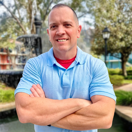 A man wearing a black cap, sunglasses, and a dark shirt with a logo, standing outdoors with his hand over his chest, surrounded by a group of blurred people.