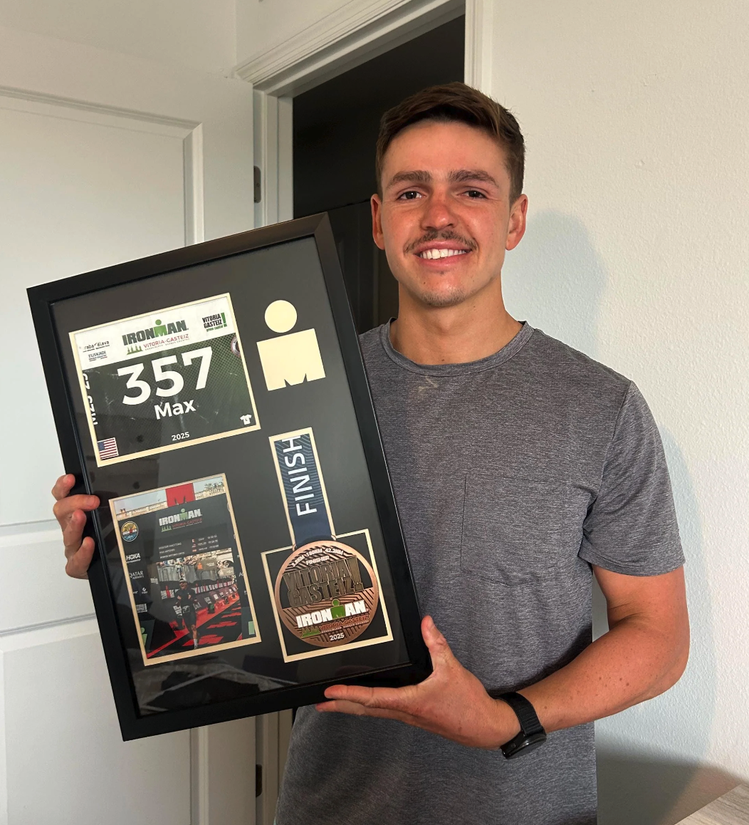 A young man in a gray t-shirt smiling and holding a framed Ironman race memorabilia, including a bib, finisher medals, and photos from the event.