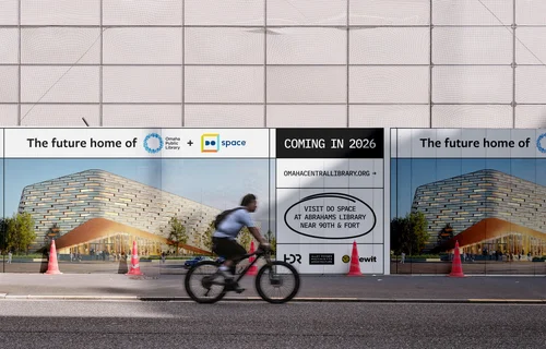 Construction fence wrapped in Omaha Central Library campaign graphics, a cyclist passing