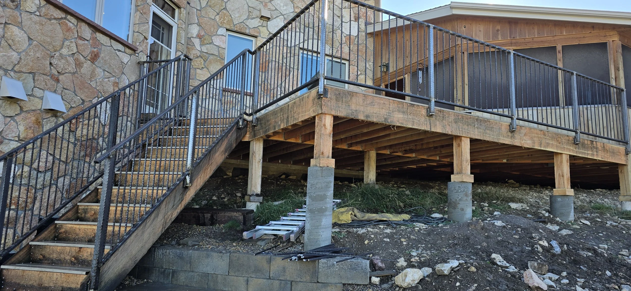 Construction site of a wooden deck with black metal railing, attached to a house with stone exterior. The deck is supported by wooden beams and concrete piers, with construction materials and tools underneath.
