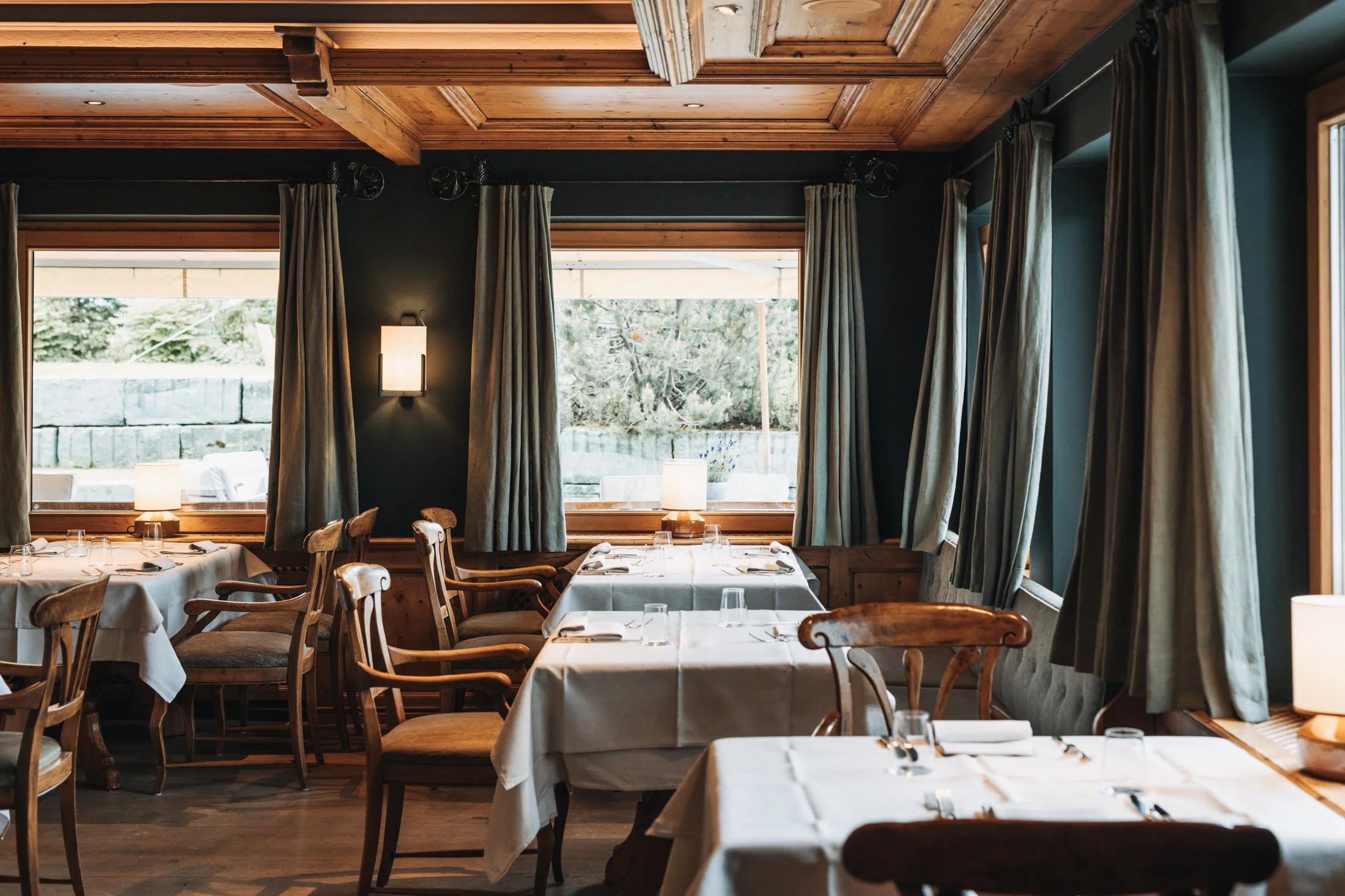 Gedeckte Mittagstische mit Blick aus den Fenstern in den Berghofgarten. Dunkle, gedeckte Wand- und Stofffarben dominieren die elegante Szene.