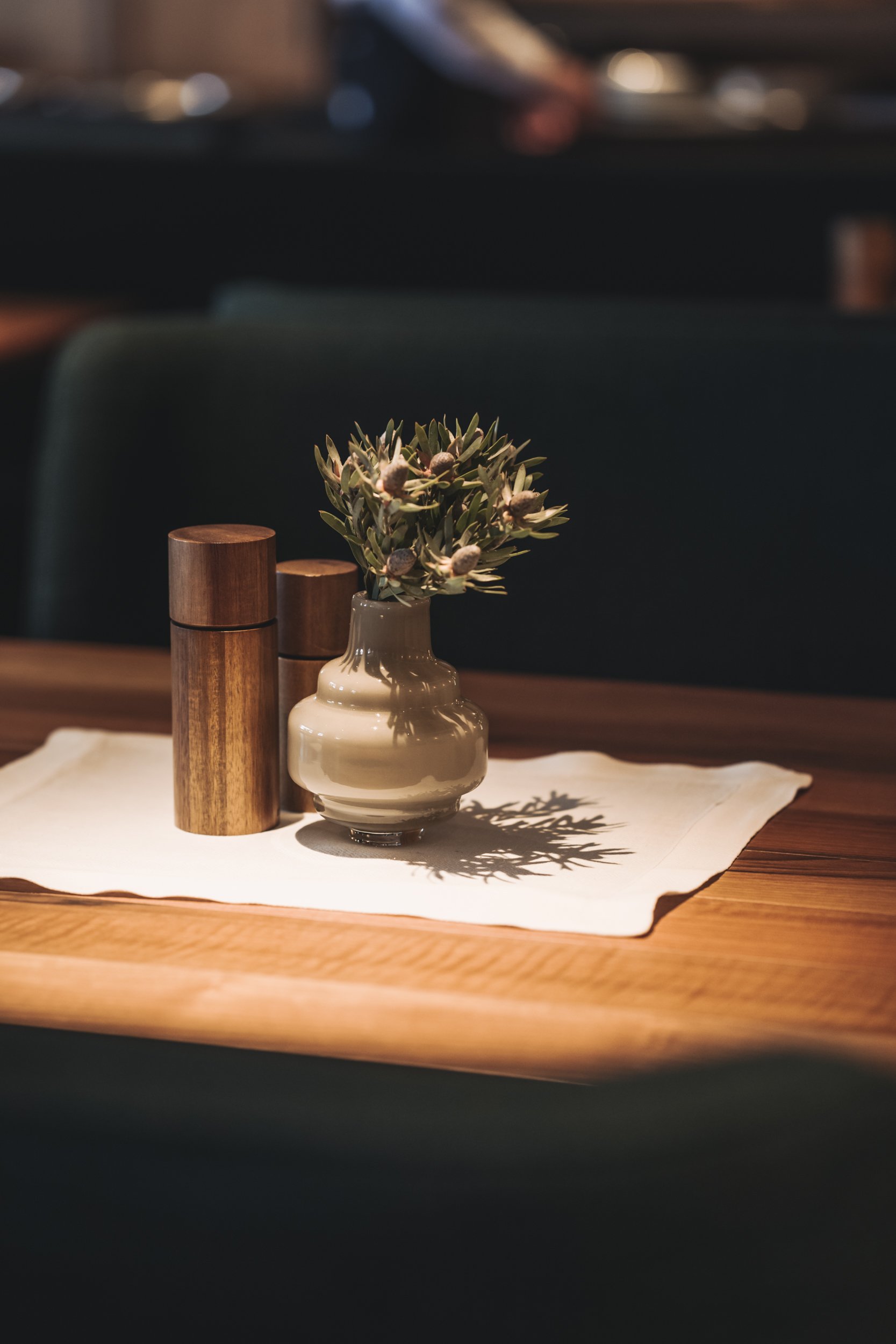 Hölzerene Salz-/Pfefferstreuer auf einem Leinenset, daneben ein kleiner alpiner Blumengruß in einer handwerklich gearbeiteten Tonvase.