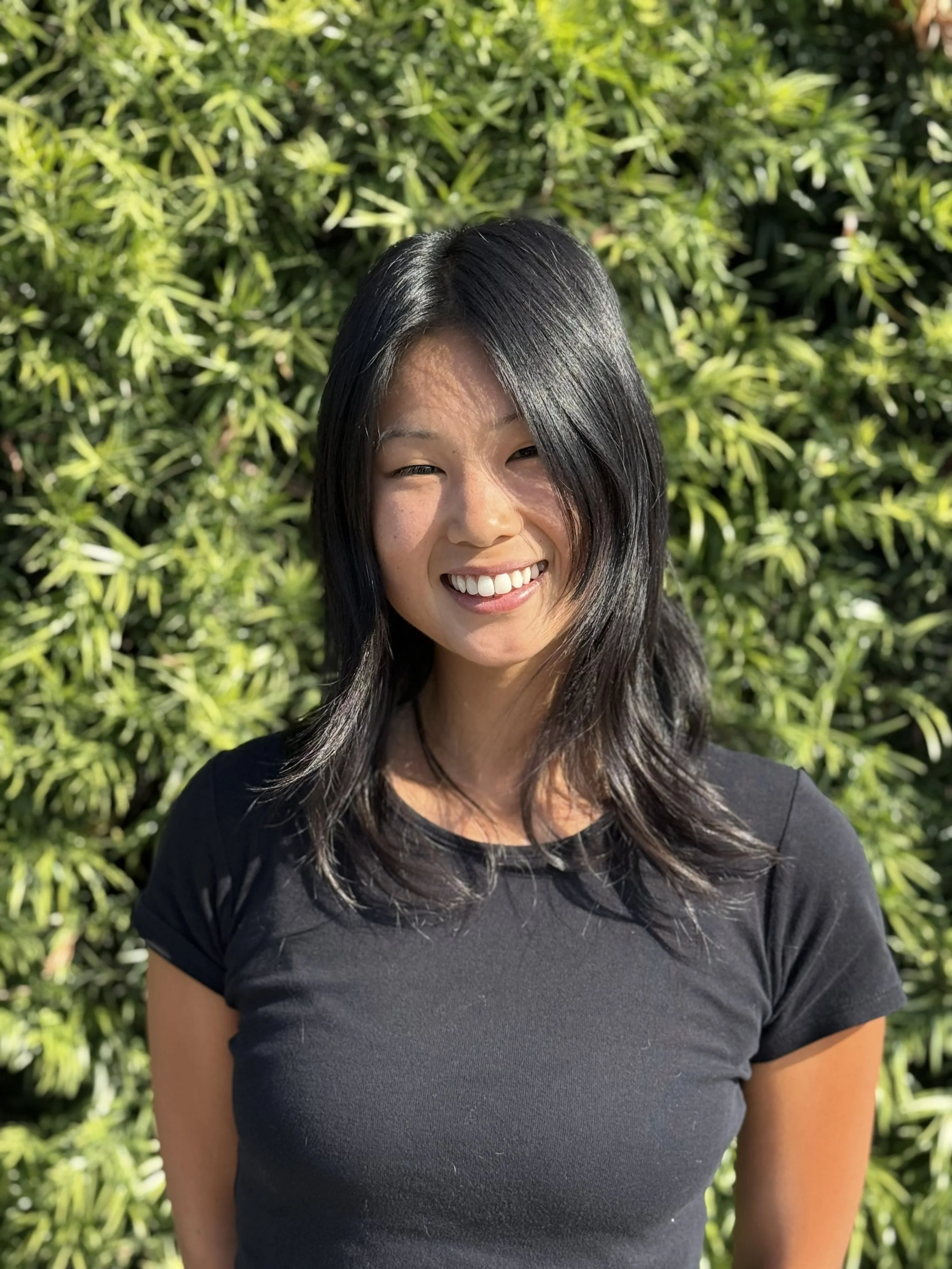 A woman smiling and standing outdoors in front of a brick wall covered with autumn-colored ivy leaves.