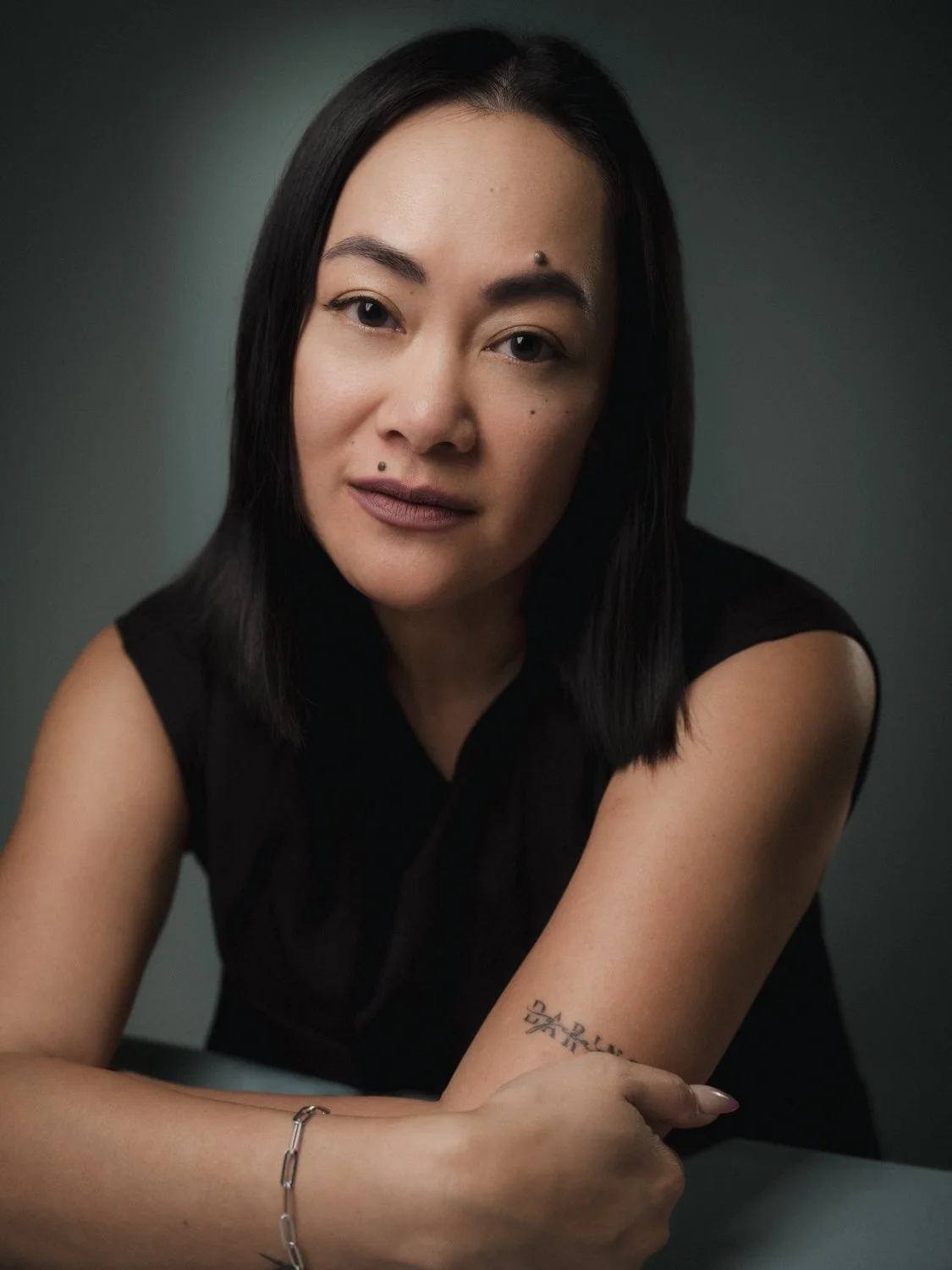 A woman with black hair and multiple facial piercings, wearing a black sleeveless top, sitting with her arms crossed on a gray surface against a dark background.
