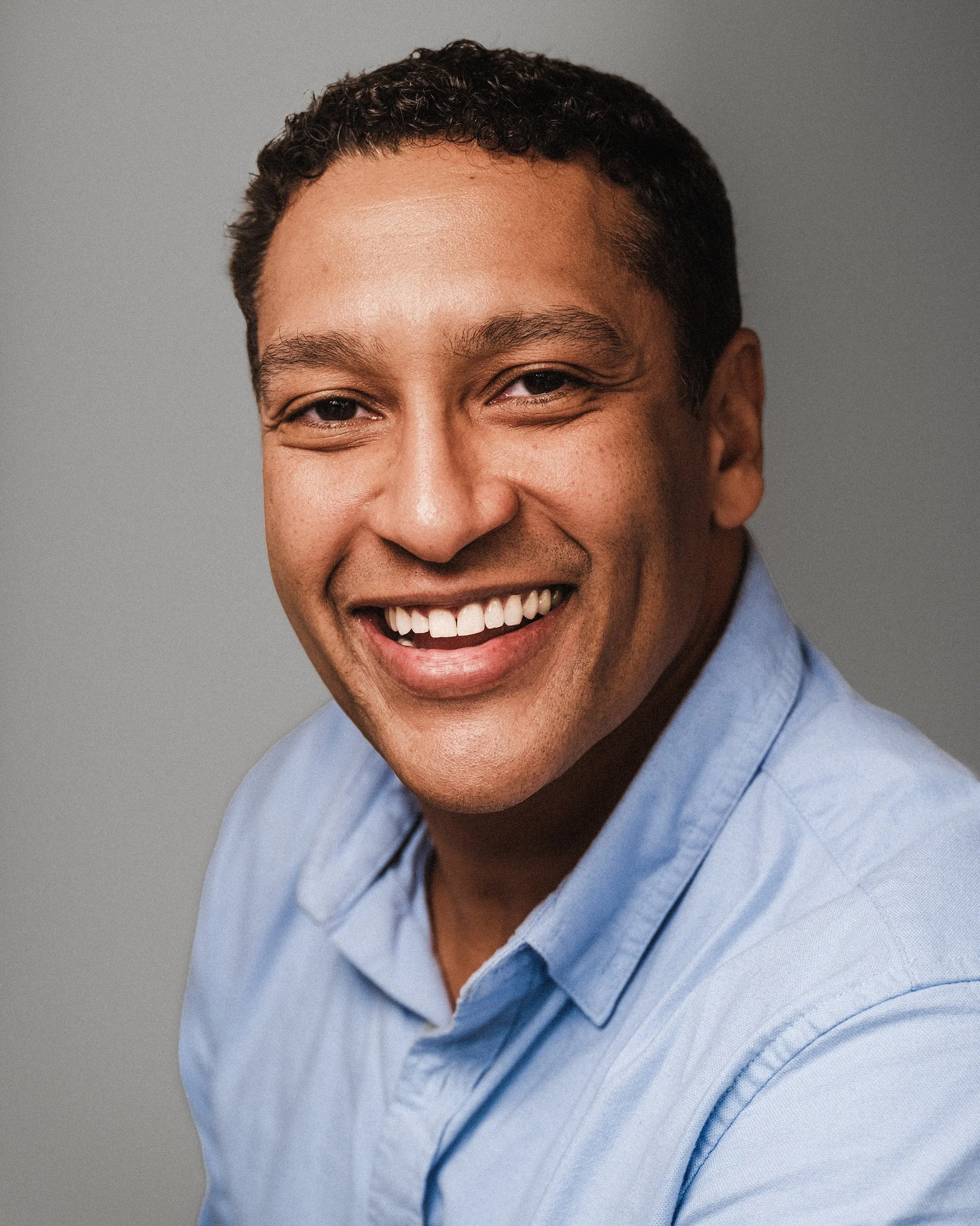 Close-up of a smiling man wearing a light blue collared shirt against a plain gray background.