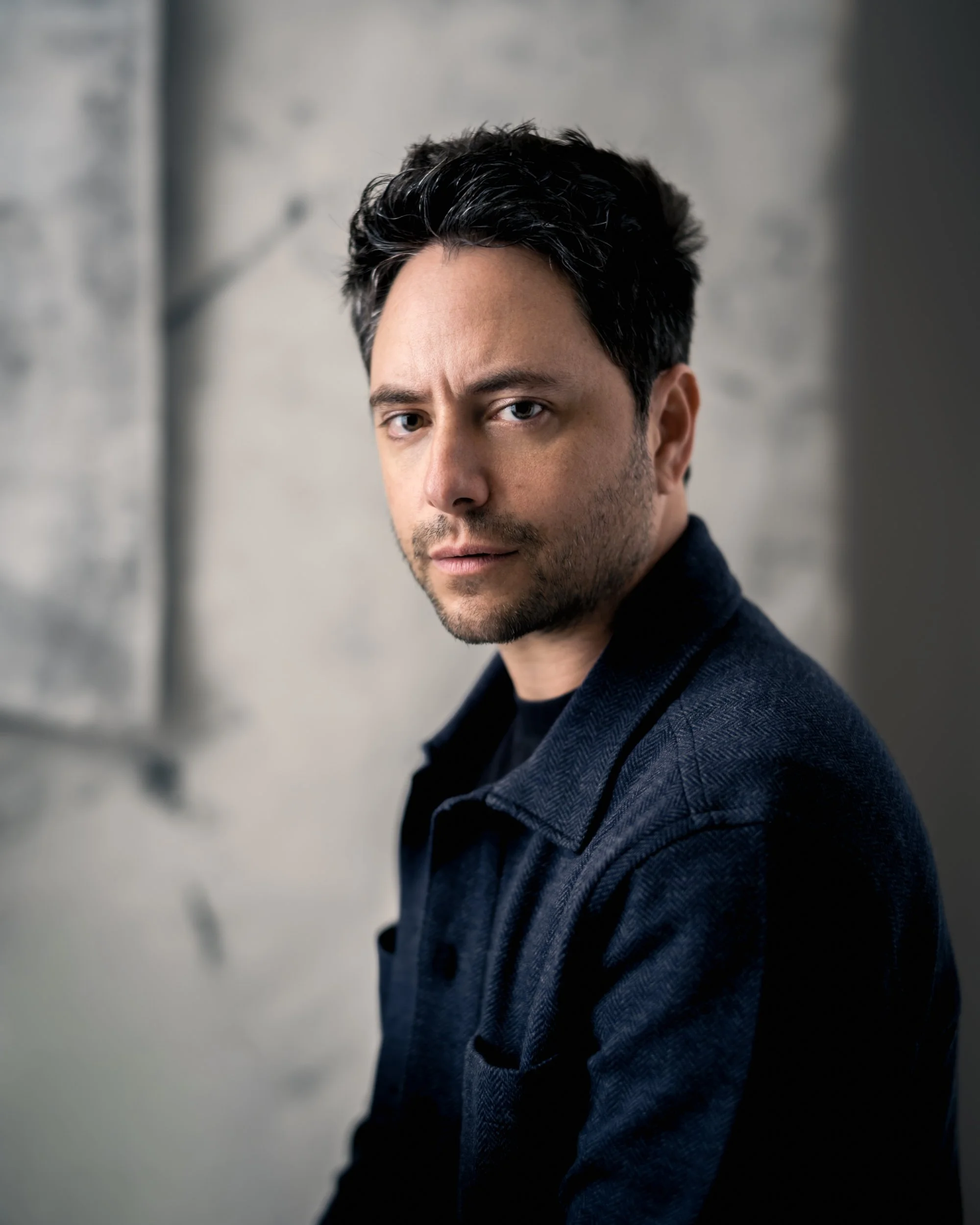 Close-up portrait of a man with dark hair and light skin, wearing a dark blazer, against a neutral background.