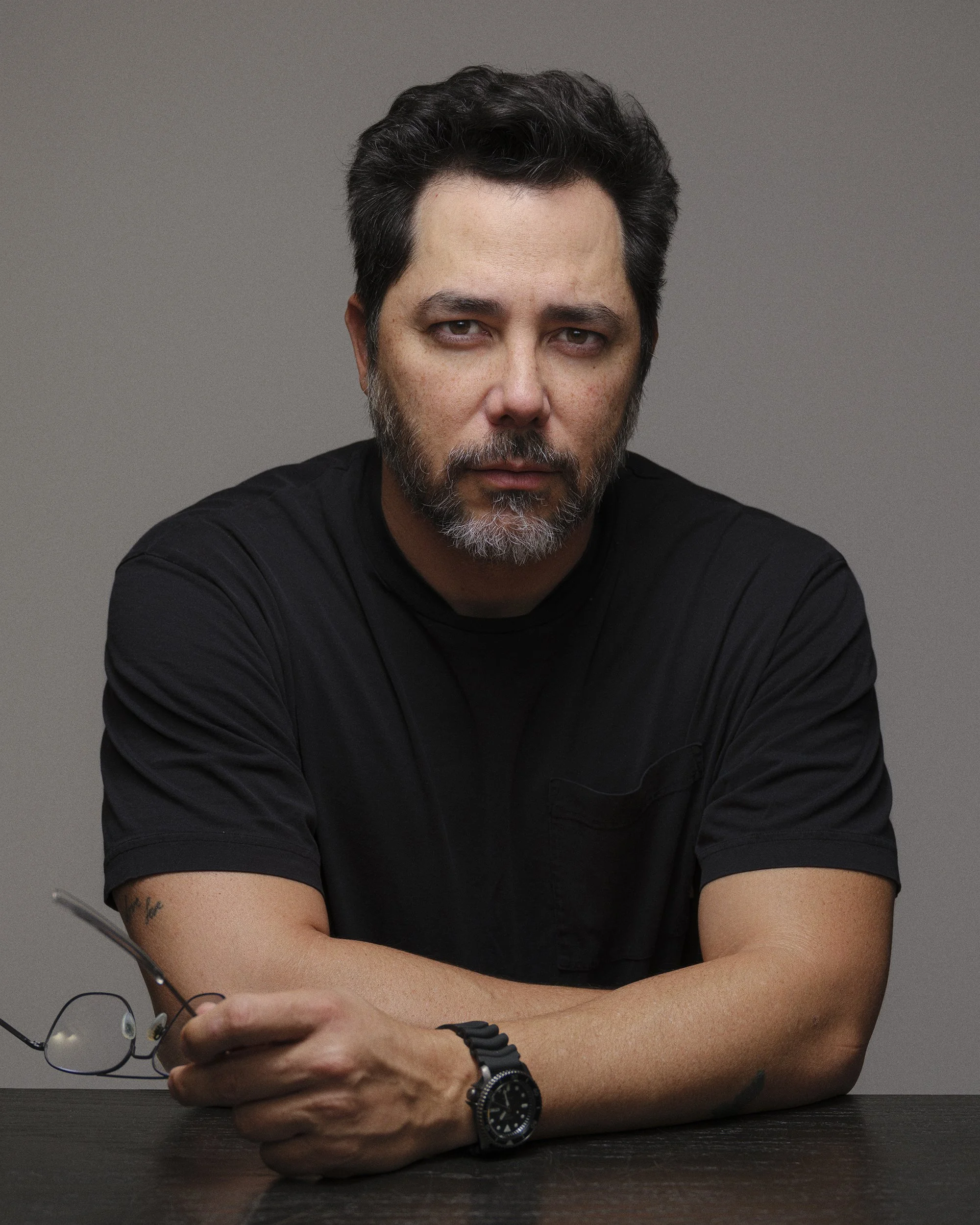 A man with dark, wavy hair, a beard, and a mustache, wearing a black shirt and a wristwatch, sitting at a dark wooden table with a gray background, holding glasses in his hand.