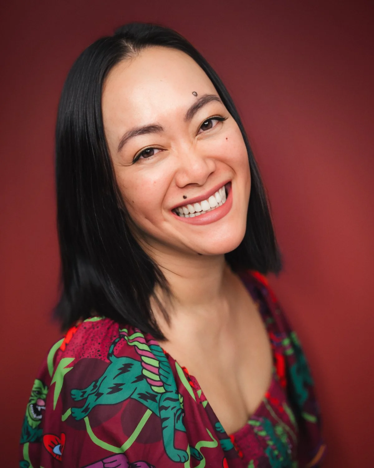 A woman with black hair smiling, wearing a colorful patterned top, standing in front of a maroon background.