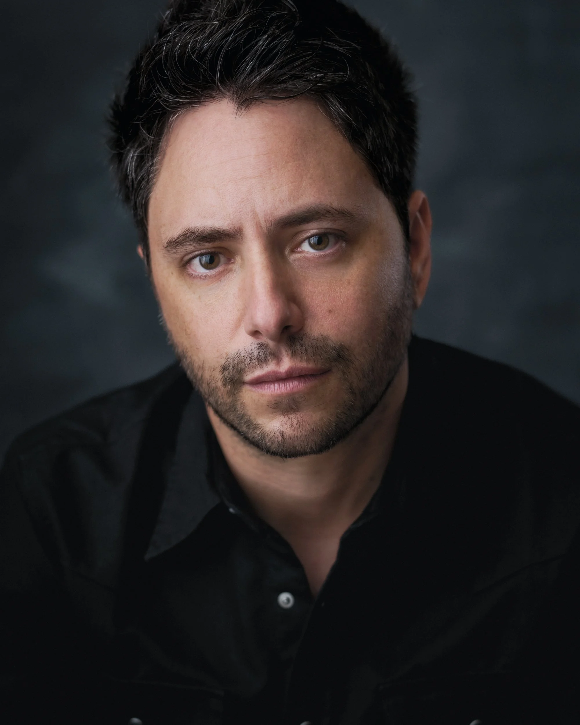 Close-up portrait of a man with dark, slightly curly hair, blue eyes, and facial hair, wearing a black shirt, against a dark background.