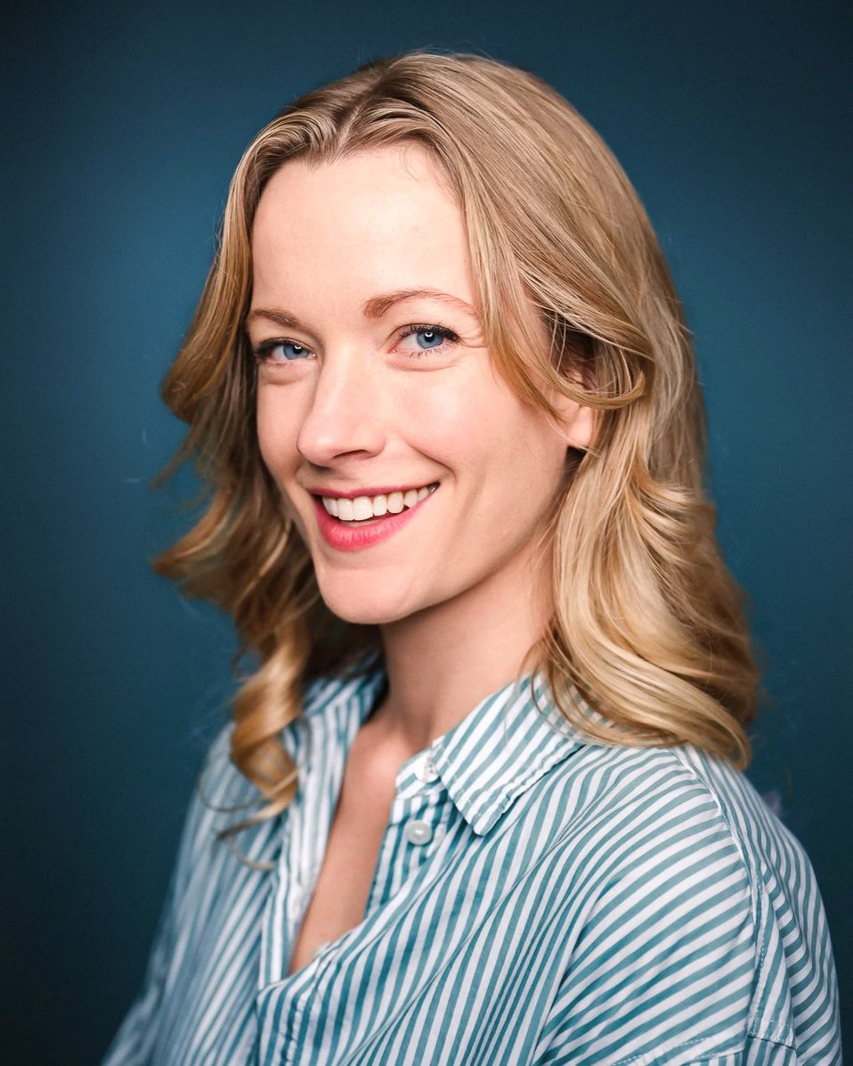A smiling woman with blonde hair and blue eyes, wearing a striped button-up shirt, against a dark teal background.