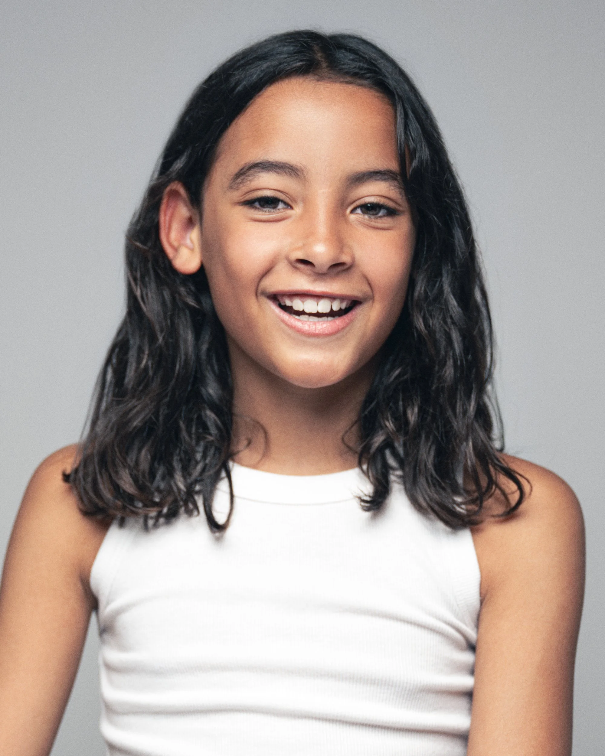 Close-up of a smiling girl with curly black hair, wearing a white tank top, against a plain light gray background.