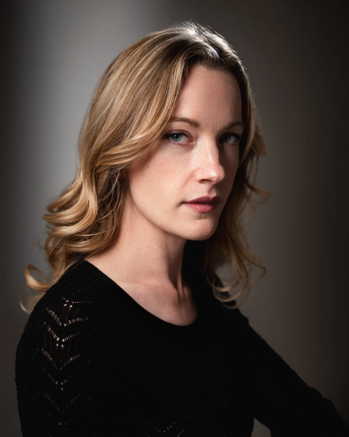 A woman with shoulder-length blonde hair, blue eyes, wearing a black top, looking at the camera with a neutral expression against a dark background.
