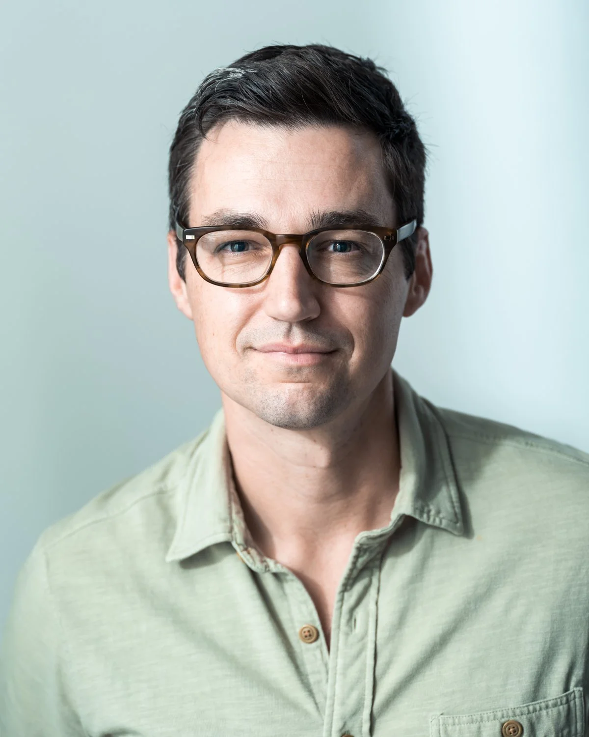 A young man with dark hair and glasses, wearing a light green shirt, looking at the camera with a slight smile.