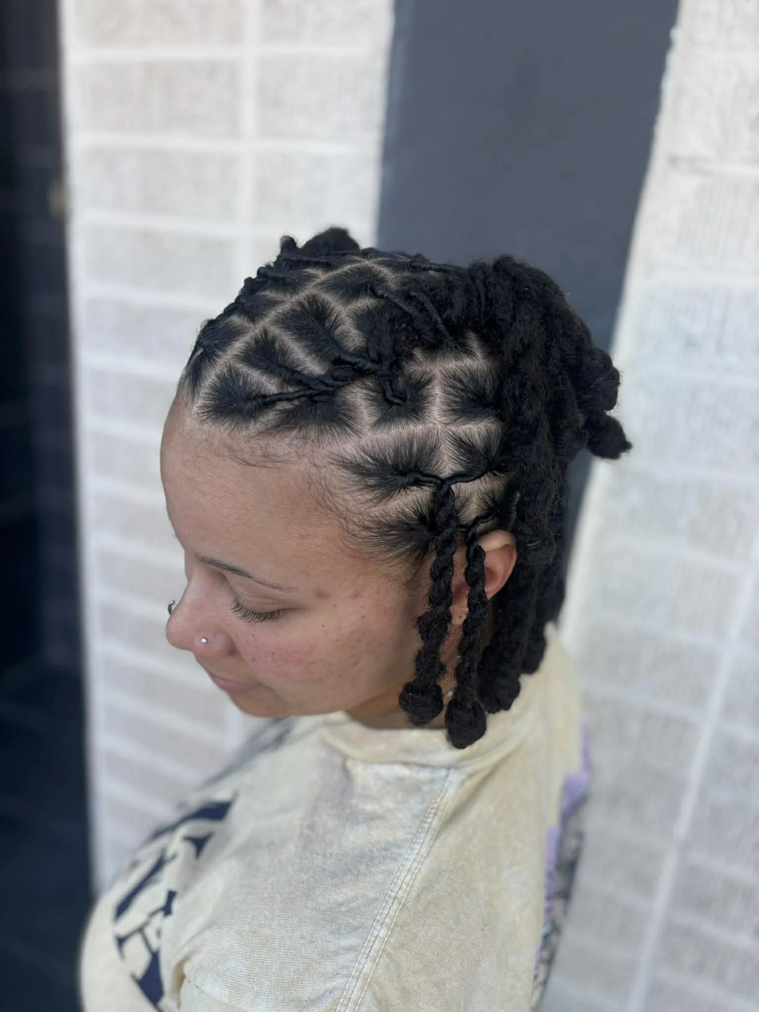 A woman with black, twisted braids and a nose piercing, standing with her head tilted downward against a gray and white brick wall background.
