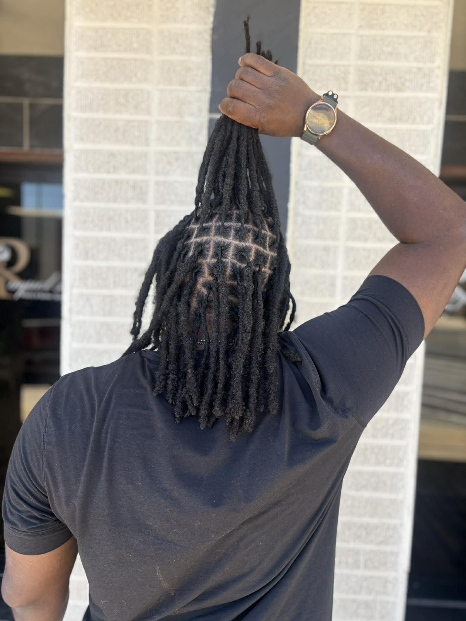 Back of a person with long locs wearing a black shirt, holding their dreadlocks up with one hand, and wearing a watch on their wrist.