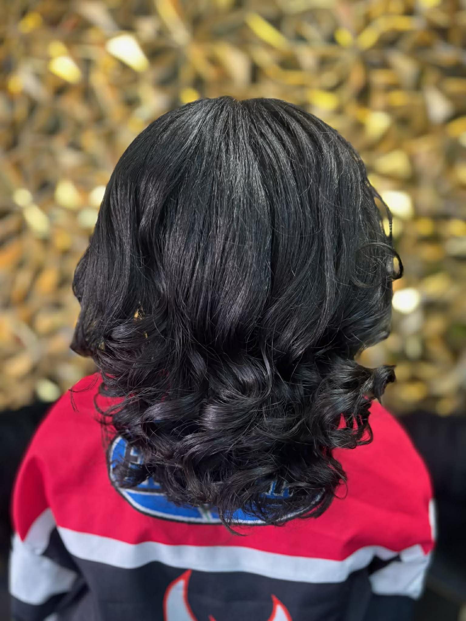 Back of a person's head with long, black, wavy hair, wearing a red and black jacket, with a blurred gold background.