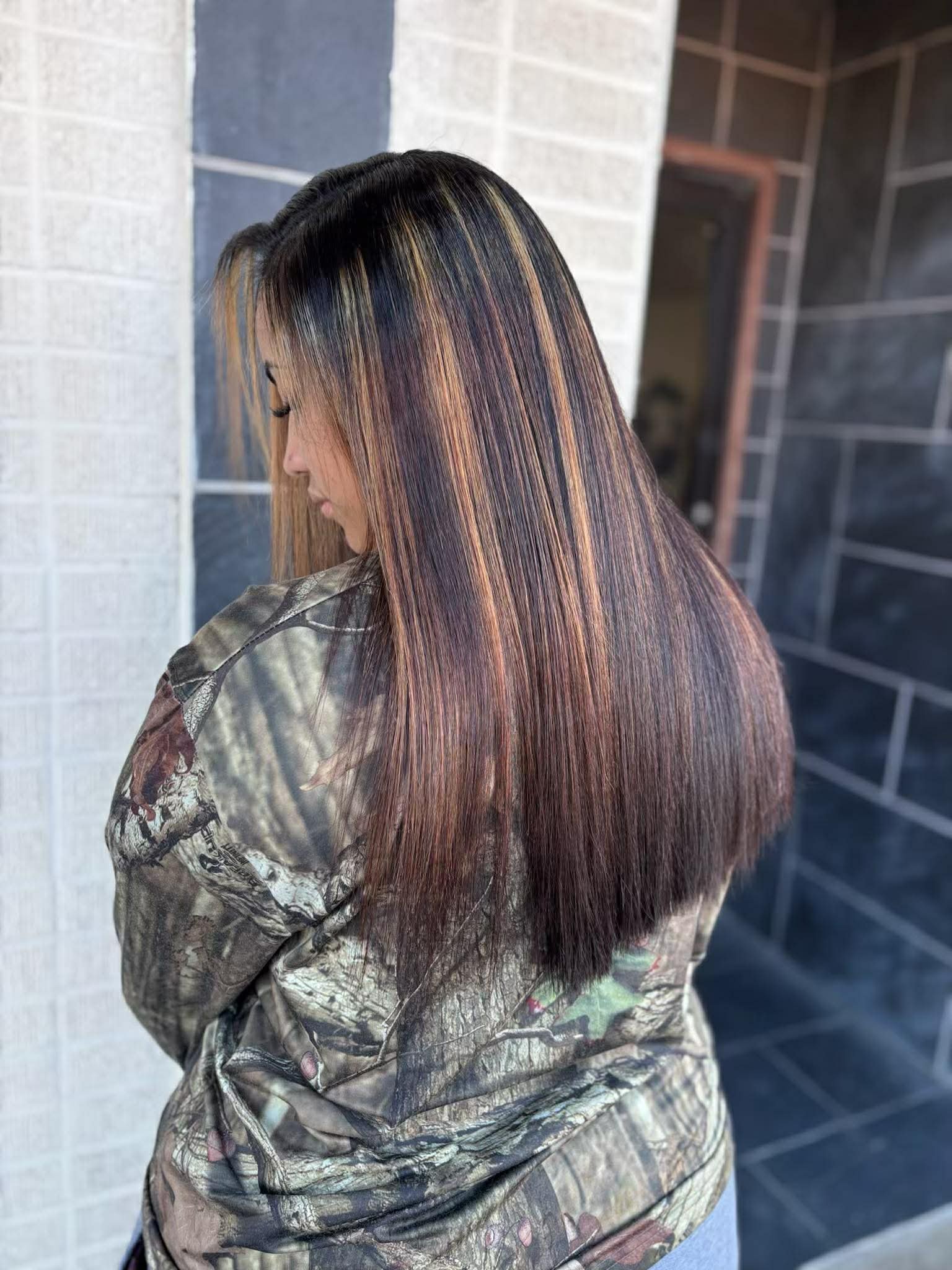 A woman with straight, shiny hair featuring blonde highlights, wearing a camouflage jacket, is looking down with her face turned to the side, standing near a tiled wall.