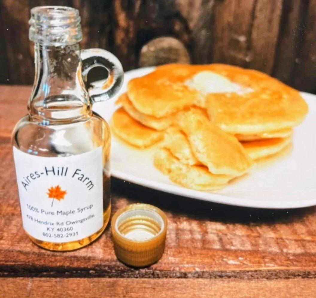 A small glass bottle of pure maple syrup with a gold cap, some pancakes on a white plate, and a wooden background.