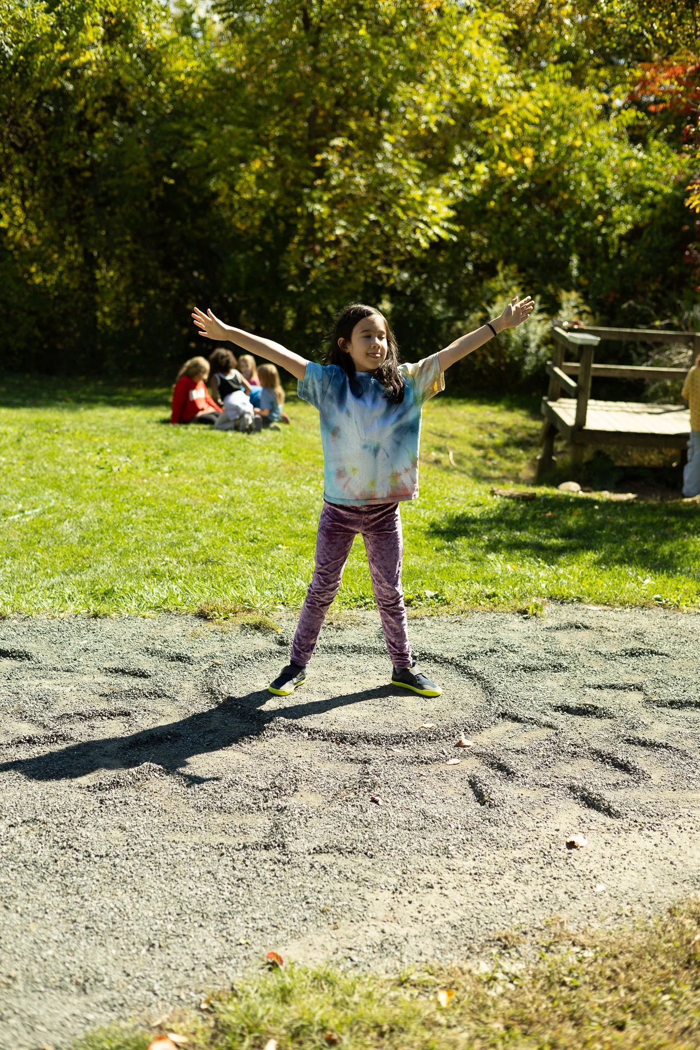 A joyful Common School student stretches their arms wide outdoors on a sunny day, surrounded by trees and open fields on 140 acres of conservation land in Amherst, MA, where children learn, play and explore the natural world.