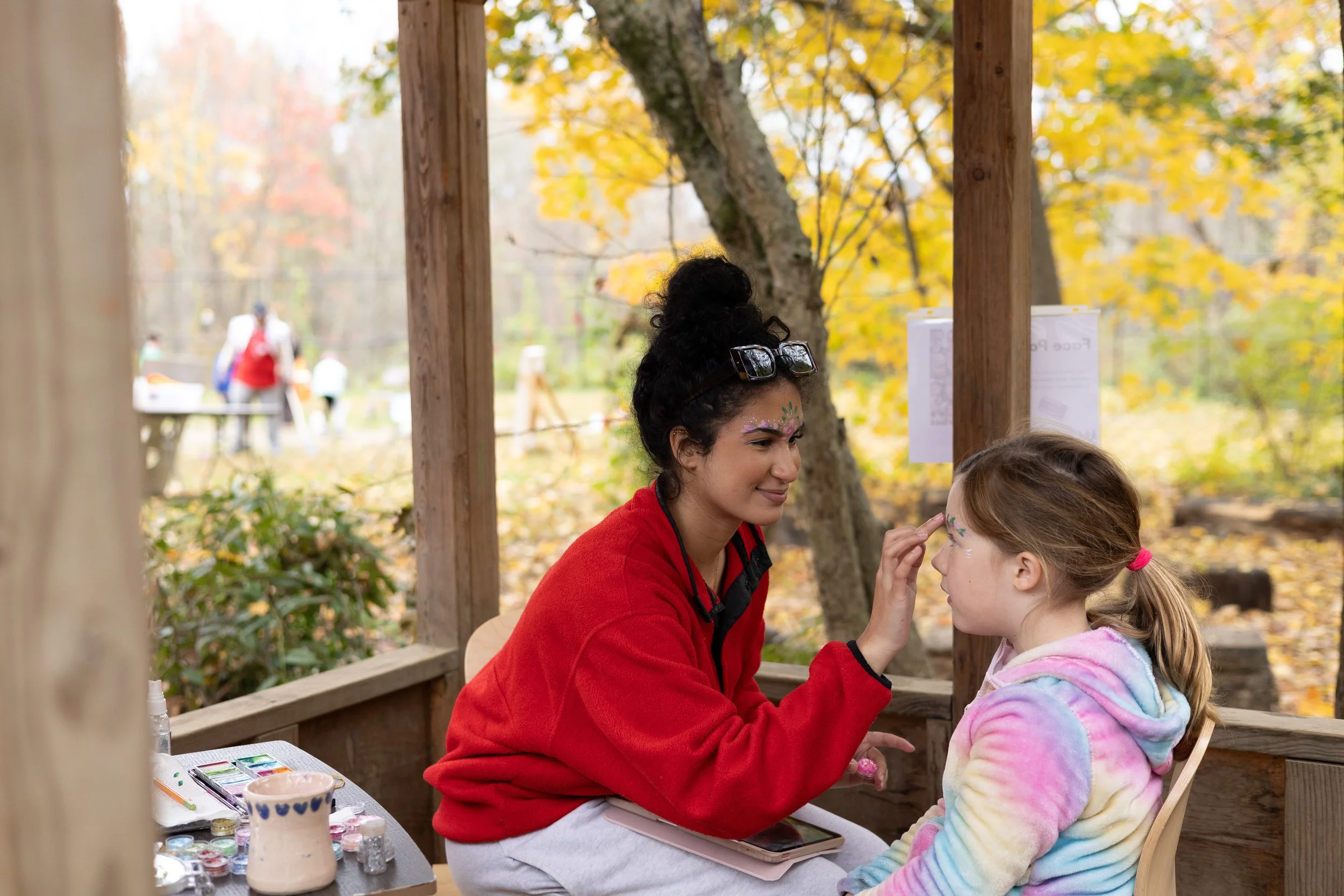 A woman with dark curly hair and face paint is sitting outside at a wooden table, painting a design on a young girl’s face, in a park during fall with yellow and orange leaves on the trees. The woman is wearing a red jacket, and the girl has a colorful tie-dye hoodie.