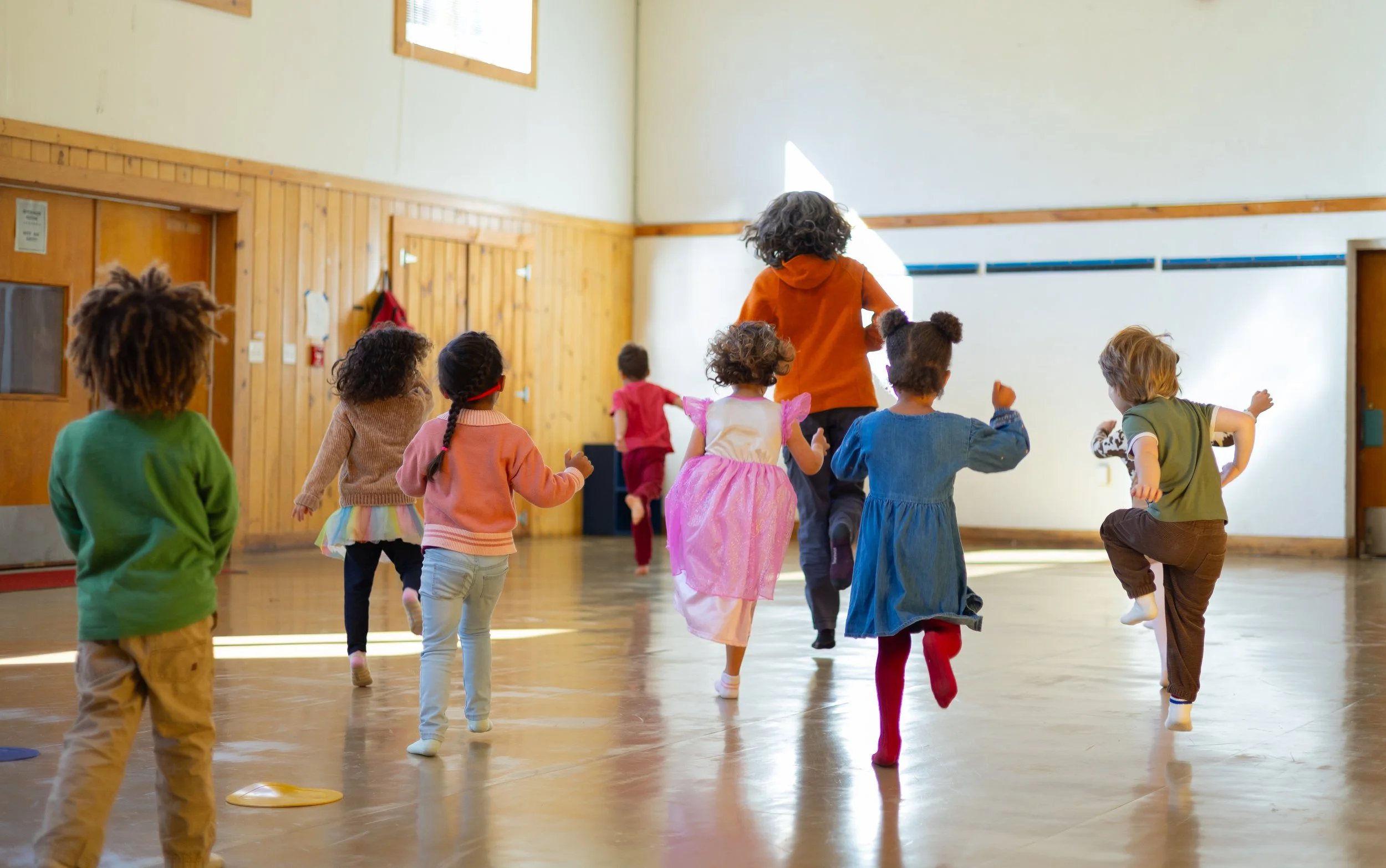 Children dressed in colorful clothing and costumes running and playing inside a gymnasium or large indoor space with wooden paneling on the walls.