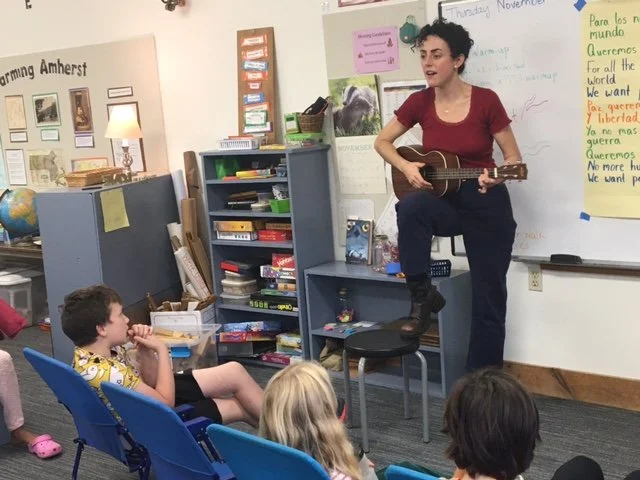 Adult with ukulele standing with one leg on a stool with mouth open in classroom, in front of kids with their backs to the camera