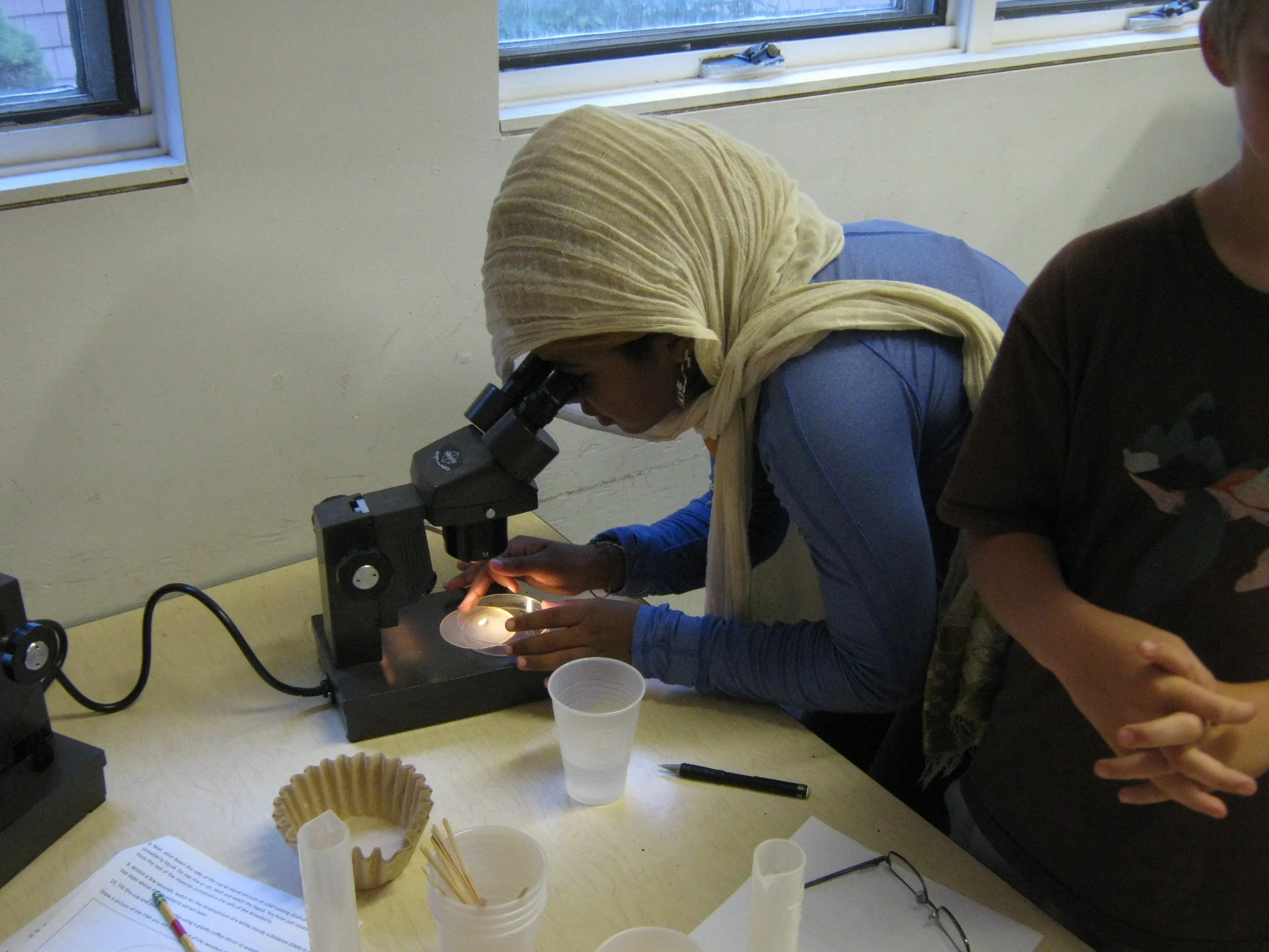 elementary-age student in a white hijab and blue shirt looking through a microscope