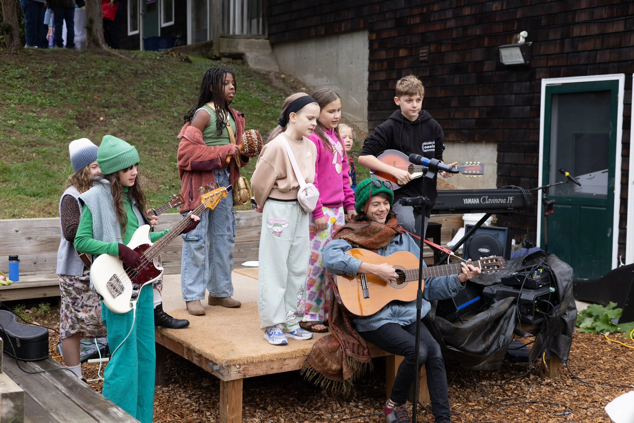 Children and adults performing outdoors on a wooden stage with musical instruments like guitars and a keyboard, with a house and trees in the background.
