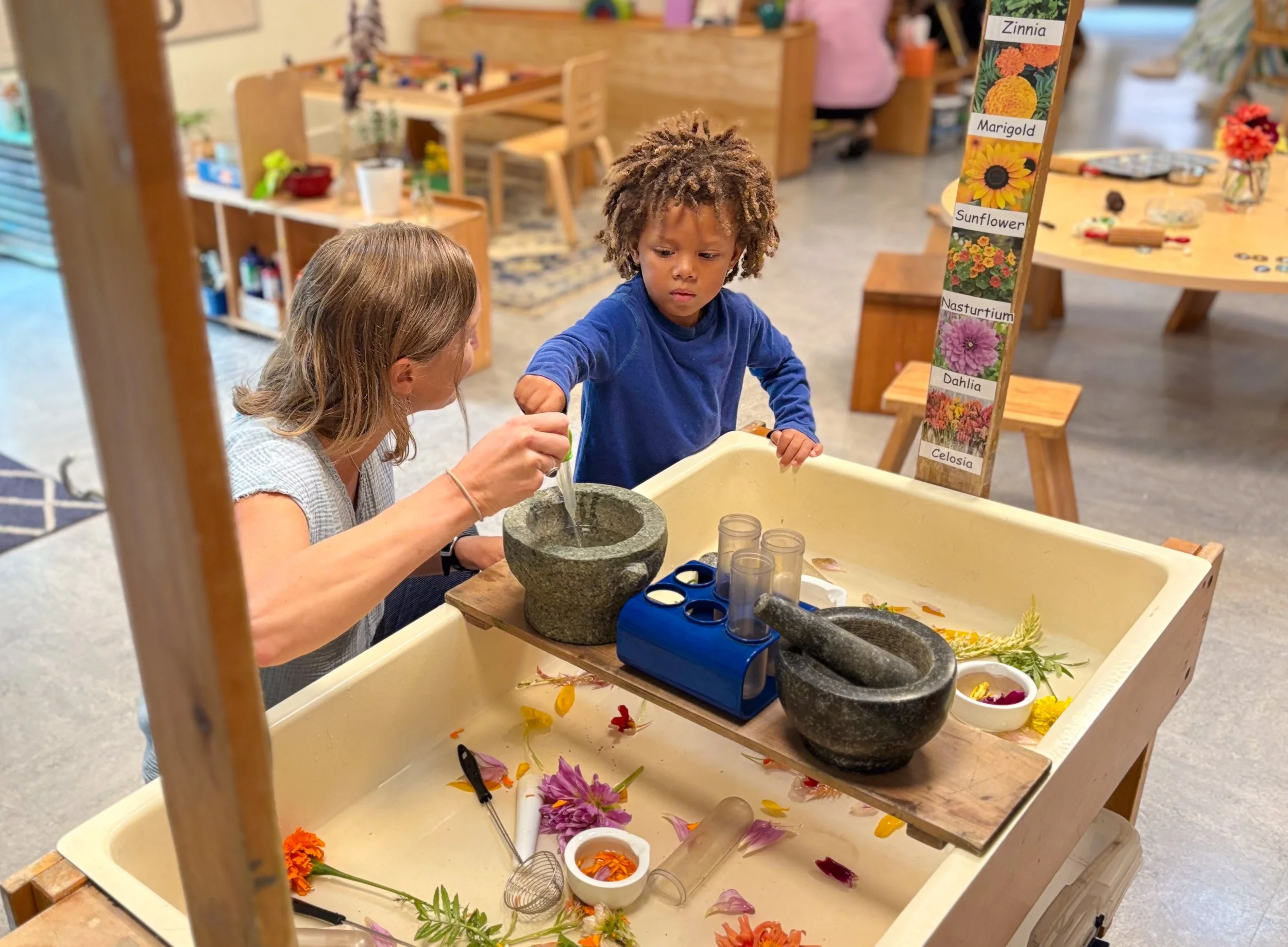 A young boy with curly hair and a blue shirt looking at a woman with long hair as they work together on a flower arrangement craft project at a table. There are various flowers, petals, and gardening tools on the table, and a colorful flower identification chart in the background.