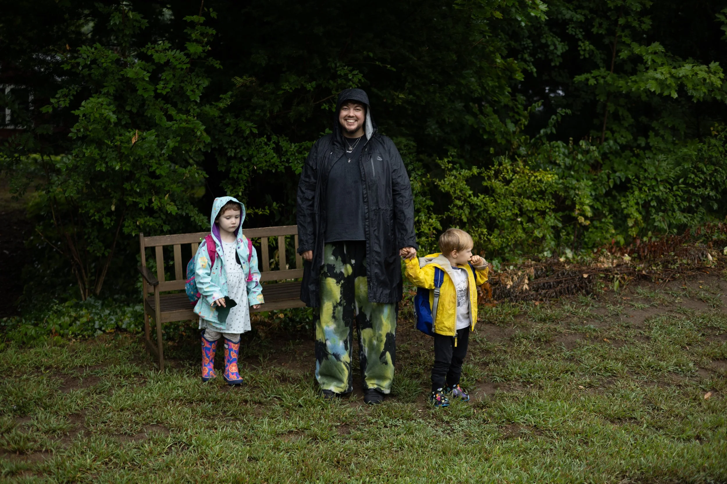 A man and two children standing outdoors in front of green foliage. The man is smiling, wearing a black rain jacket and colorful pants. The girl on the left is wearing a raincoat with a hood and rain boots, holding a small object. The boy on the right is wearing a yellow raincoat and backpack, holding hands with the man.