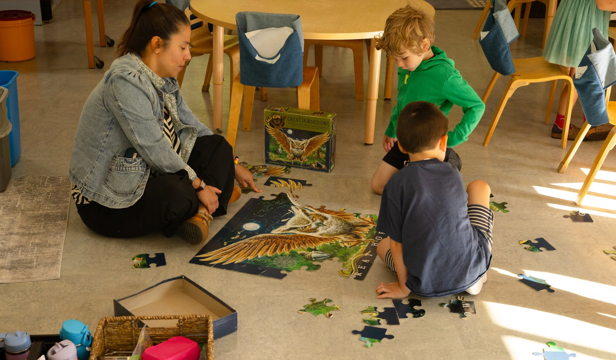 A woman and two boys sitting on the floor assembling a large jigsaw puzzle featuring a great horned owl in a science fiction or fantasy style. The scene is set in a room with wooden chairs and tables, some hanging cloth items, and shelves with various objects.