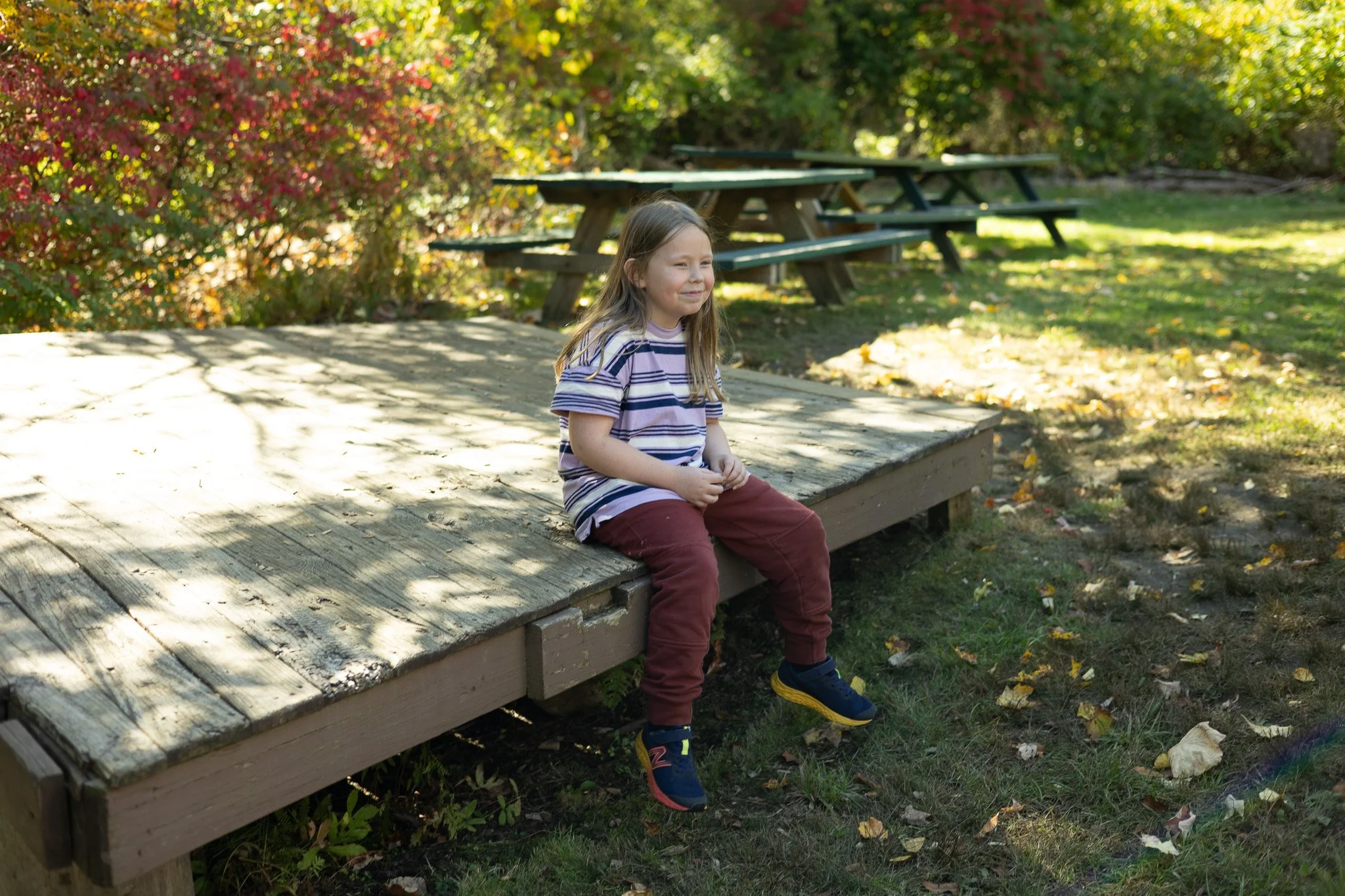 Young girl sitting on a wooden platform in a sunny park with picnic tables and autumn foliage.