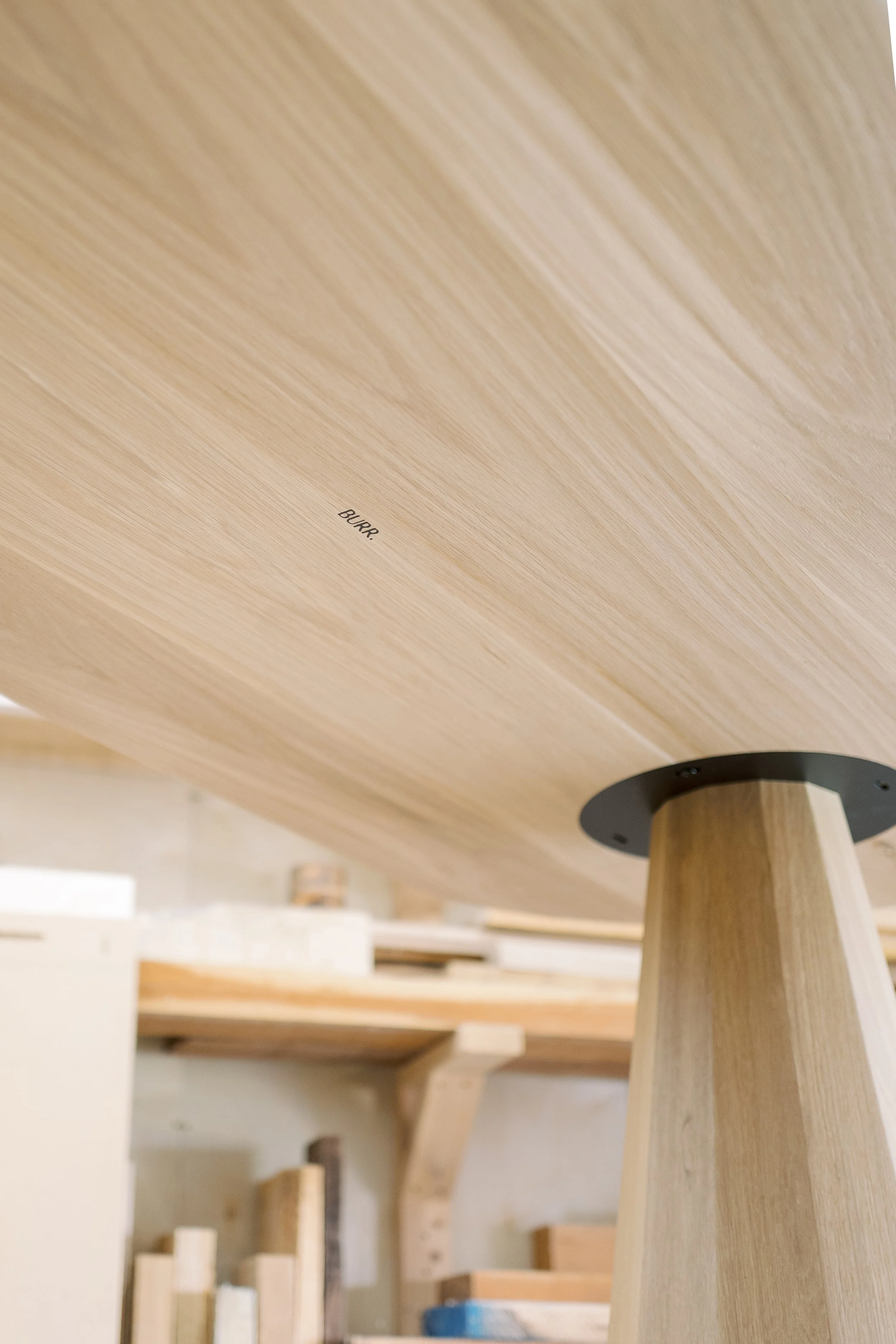 Close-up of a wooden table or piece of furniture under construction with a focus on the underside, showing a small label that reads 'BURR'. The background shows a workshop with shelves and various pieces of wood.