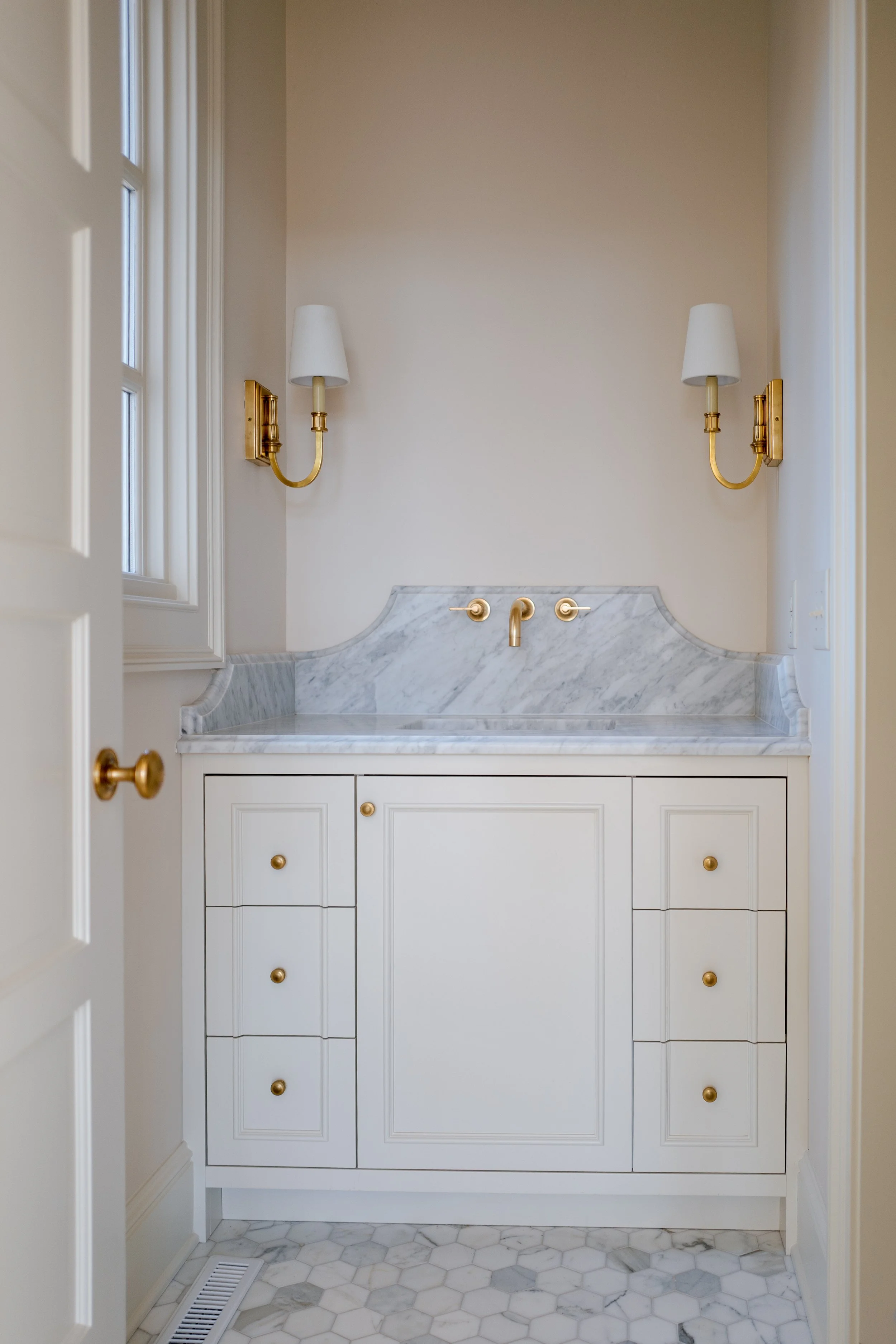 Clean bathroom vanity with white cabinets, marble countertop, brass fixtures, and two wall-mounted gold sconces with white shades.