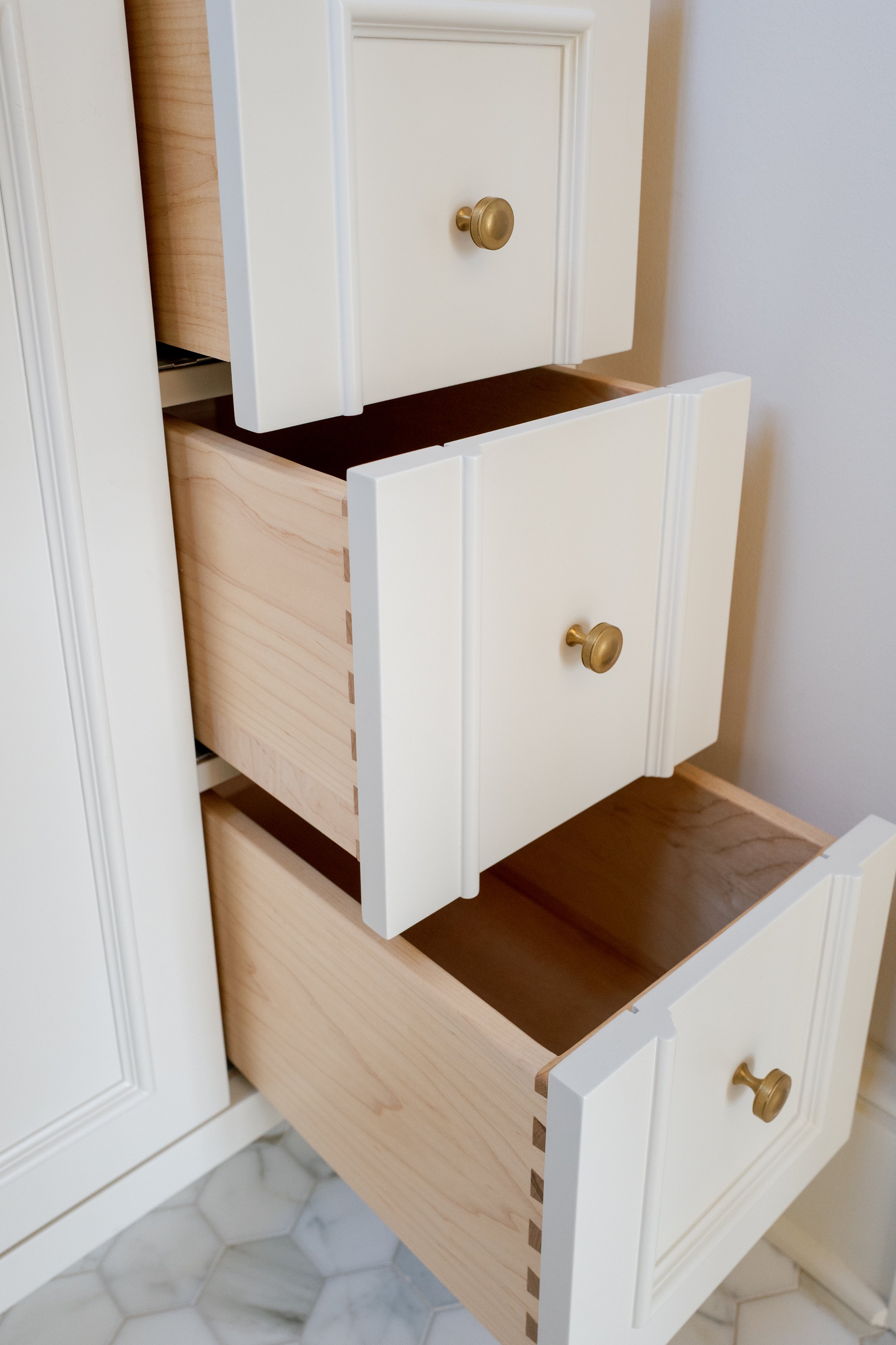 Open kitchen cabinet drawers with wooden interiors and white painted exterior, brass knobs, on a tiled floor.