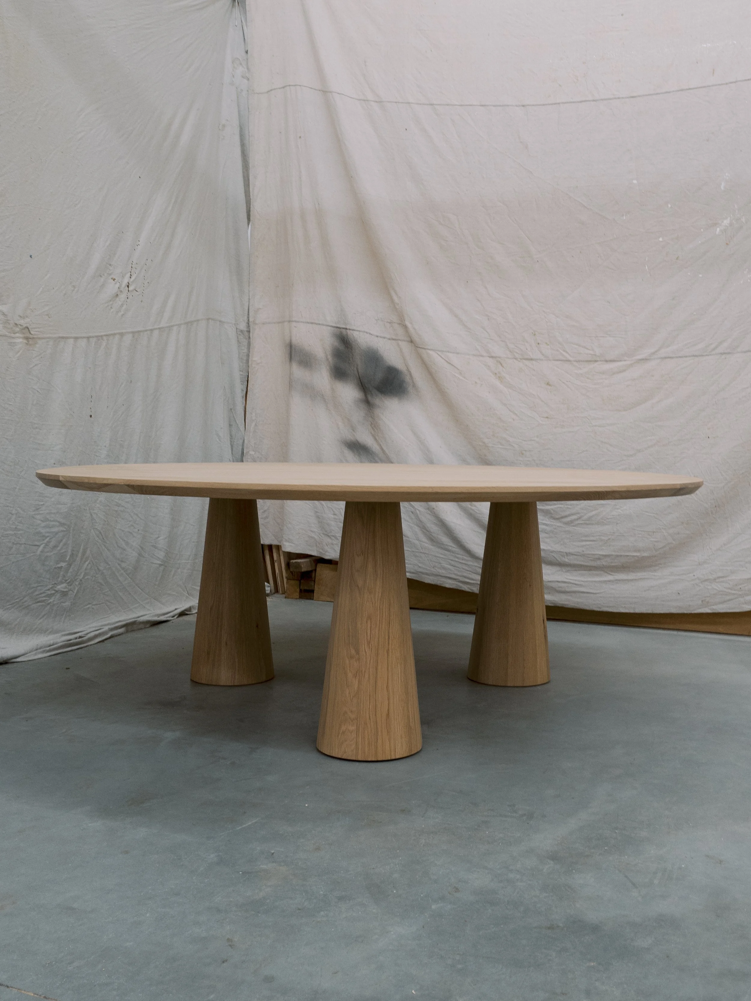 A wooden table with a smooth, flat surface and three conical legs stands on a gray concrete floor with a white fabric backdrop.