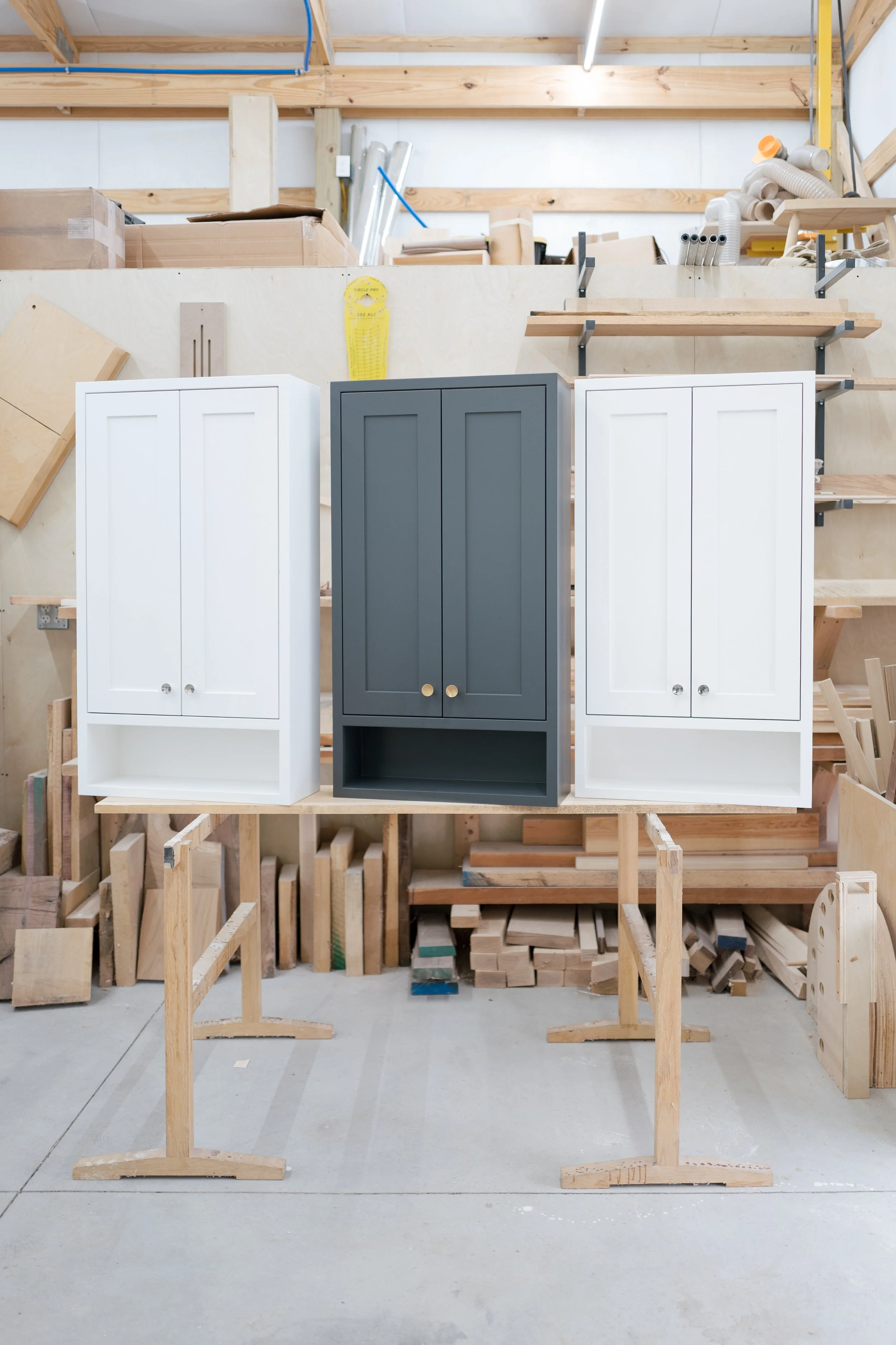 Three cabinetry pieces, two white and one dark gray, are displayed in a woodworking shop on wooden stands surrounded by various woodworking materials and tools.