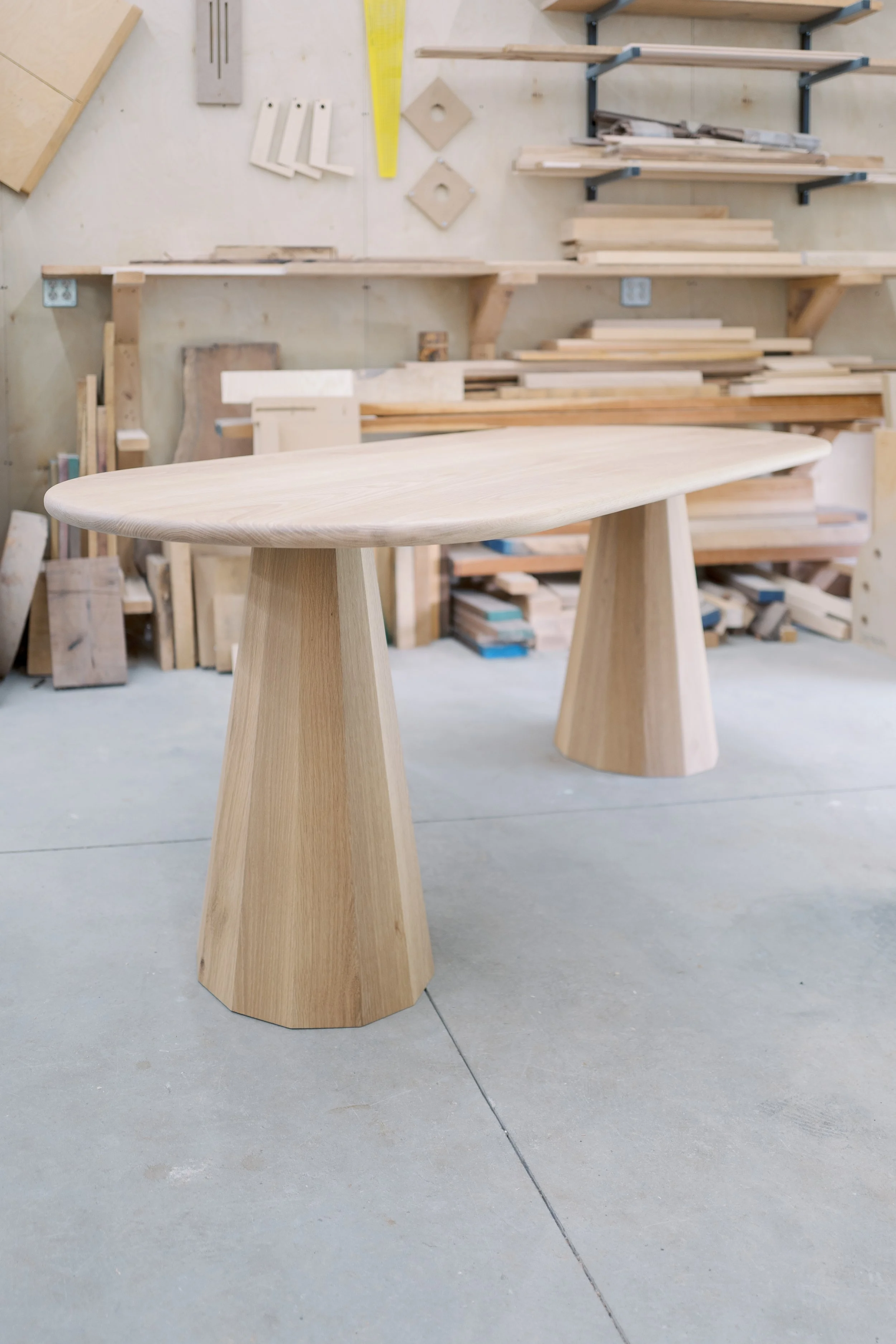 A wooden table with a smooth, rounded top and tapered, angular legs in a woodworking shop with shelves, tools, and wood pieces in the background.