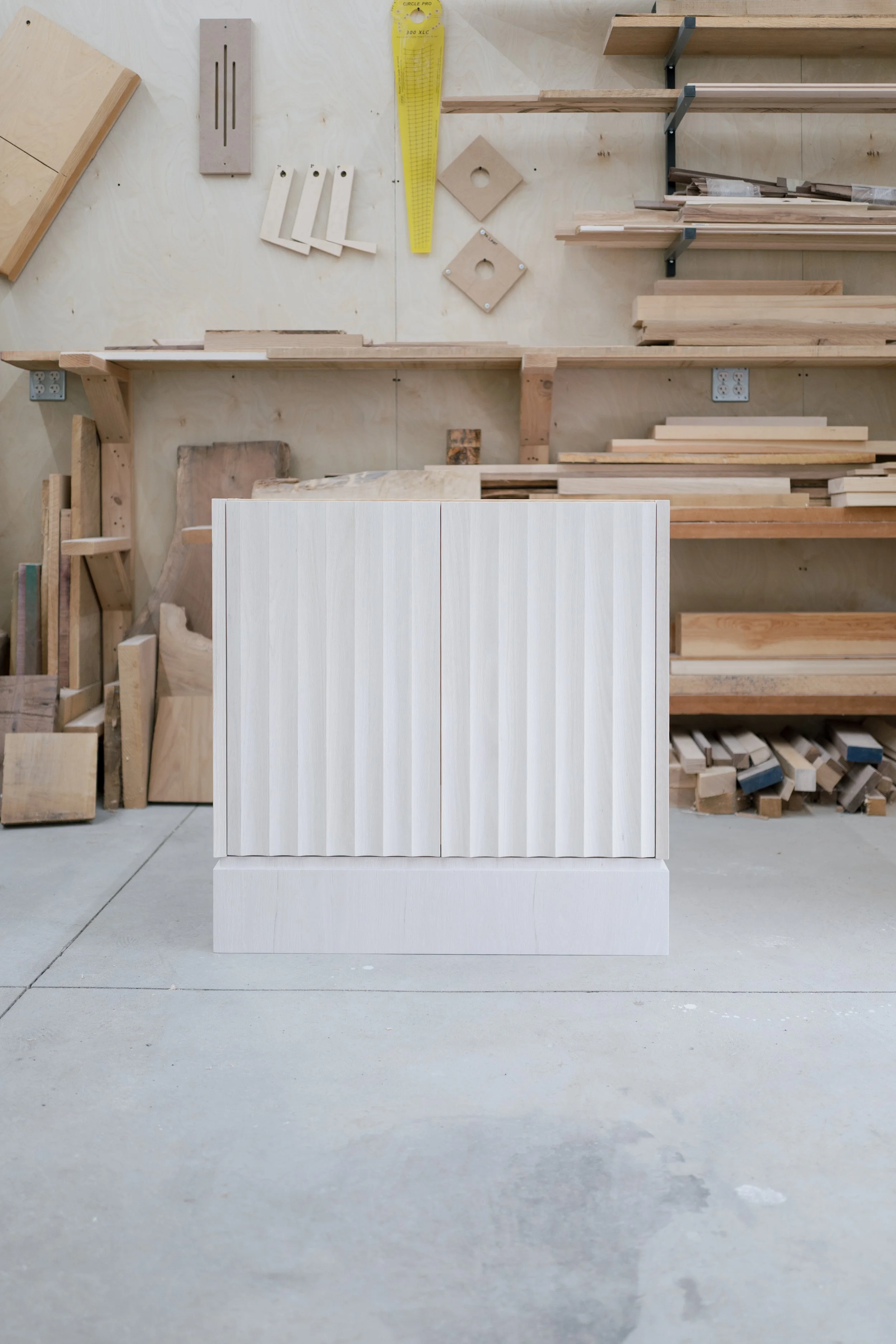 White cabinet with vertical fluting in a woodworking shop with shelves of wood and tools on the wall behind.