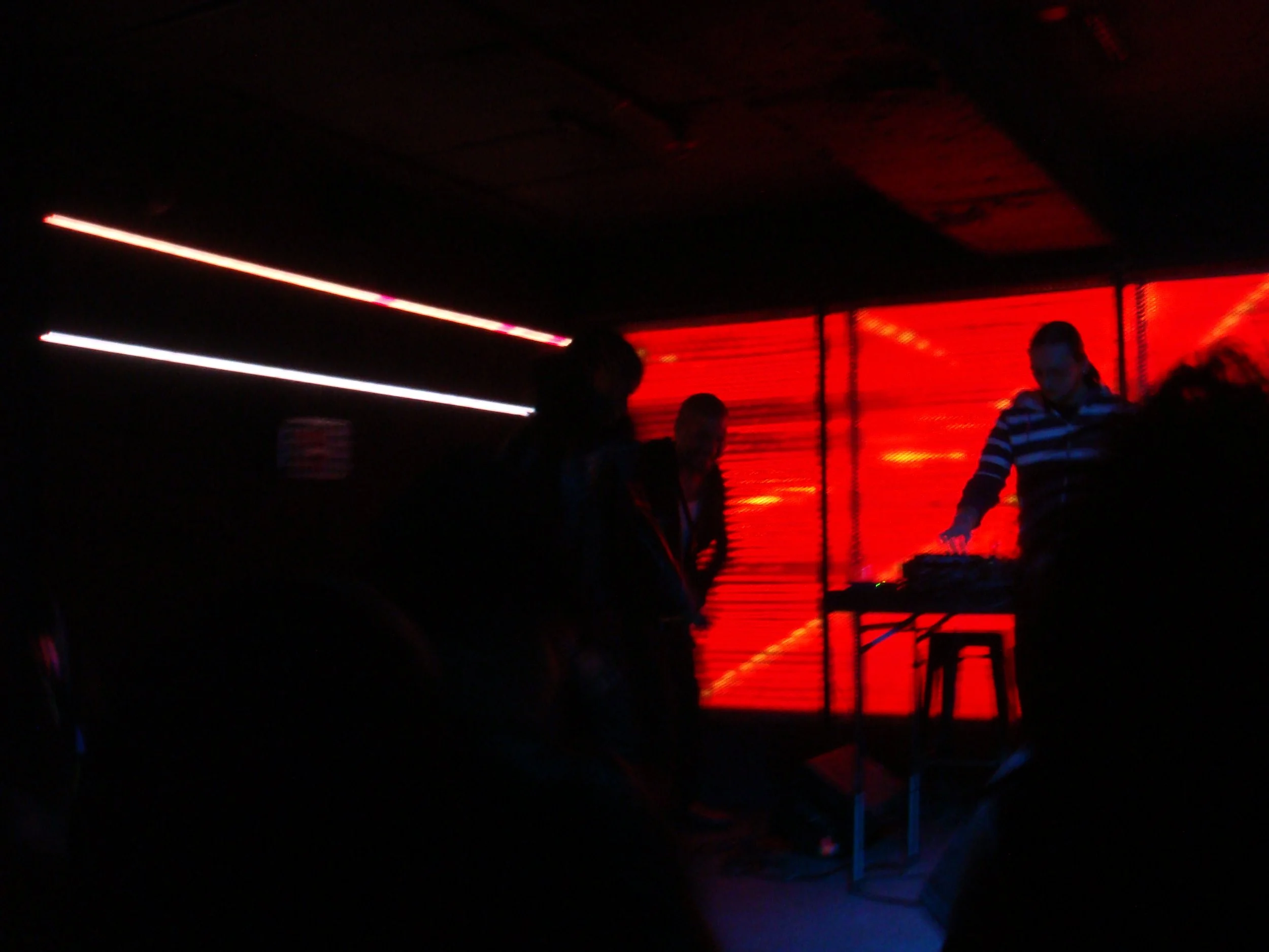 Three people in a dark room with red and white neon lights, two appear to be watching, one is operating DJ equipment.