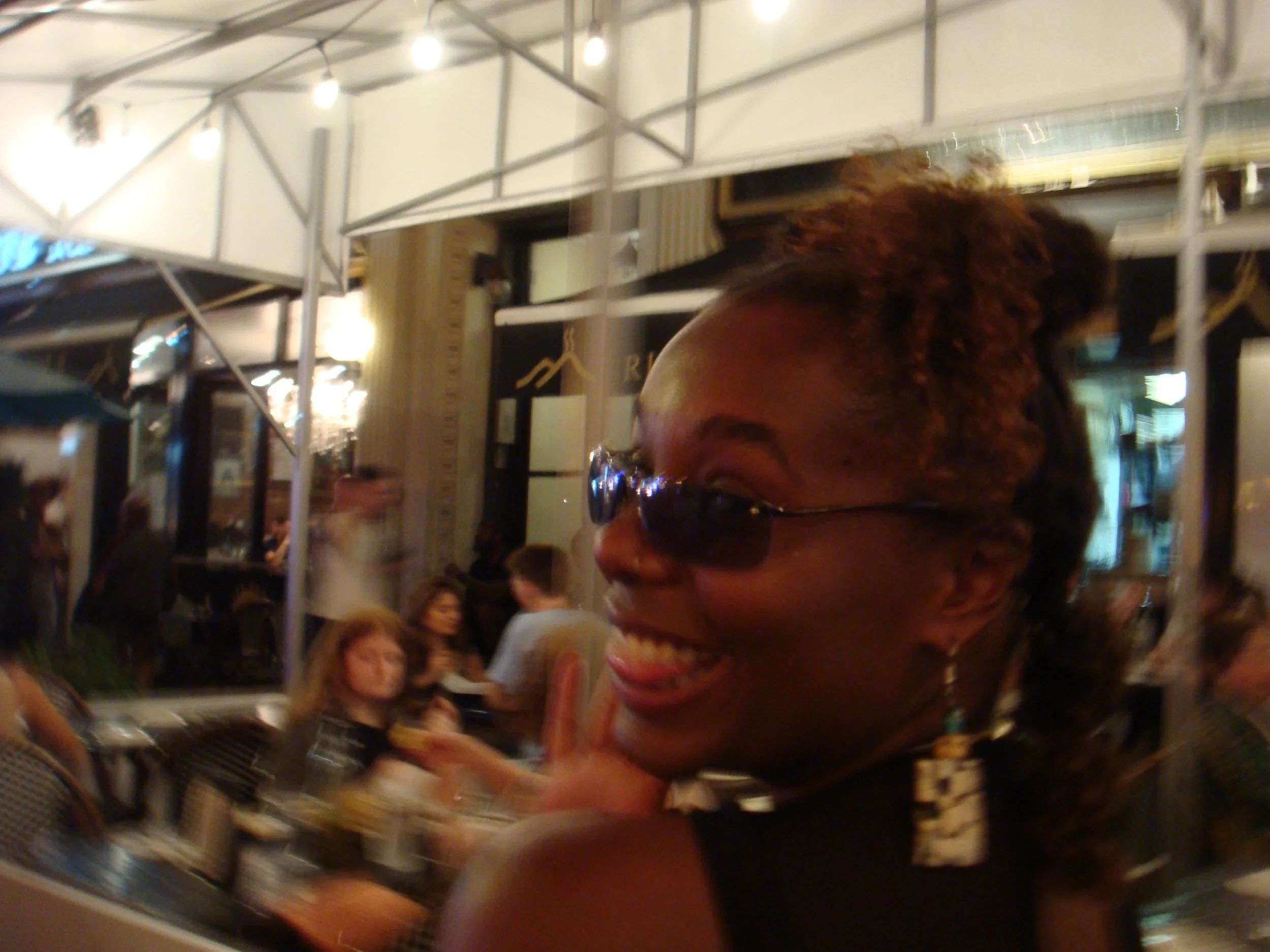 A woman smiling and wearing sunglasses at an outdoor dining area with people sitting at tables behind her.